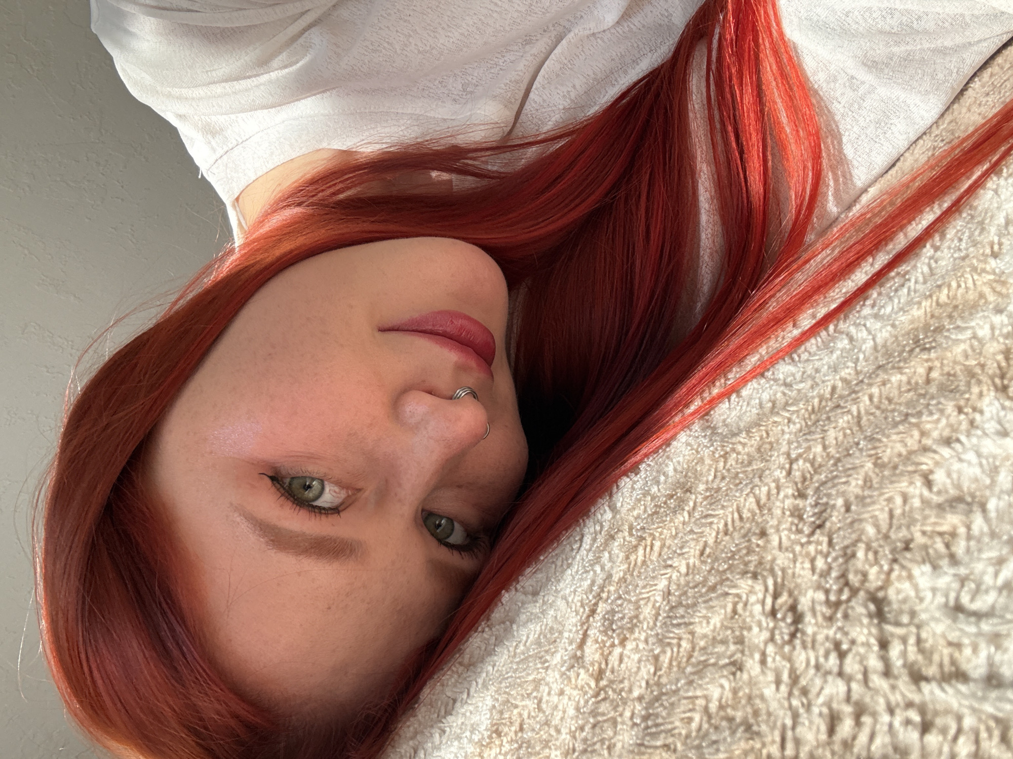 An overhead portrait features a young woman with vibrant red hair and green eyes, lying down and looking upwards towards the camera. Her head is angled to the left, resting on a soft, textured beige or cream-colored fabric, possibly a blanket or pillow, which fills the lower-right portion of the frame. Her long, fiery red hair cascades around her head and across the fabric.
She has fair skin with visible freckles and is wearing light makeup, including pink lipstick and subtle dark eyeliner accentuating her bright green eyes. A double-hoop nose ring is visible on her nose. She is wearing a light-colored, possibly white, top, the fabric of which can be seen in the upper-left of the image. The background is a plain, light grey or off-white wall, softly lit.
The lighting appears natural and soft, suggesting daytime, casting gentle shadows that define her facial features. Her expression is calm and direct. The image is composed from an elevated perspective, giving it an intimate and relaxed feel. No text is visible in the image. The location context provided is Rostov-on-Don, Russia.