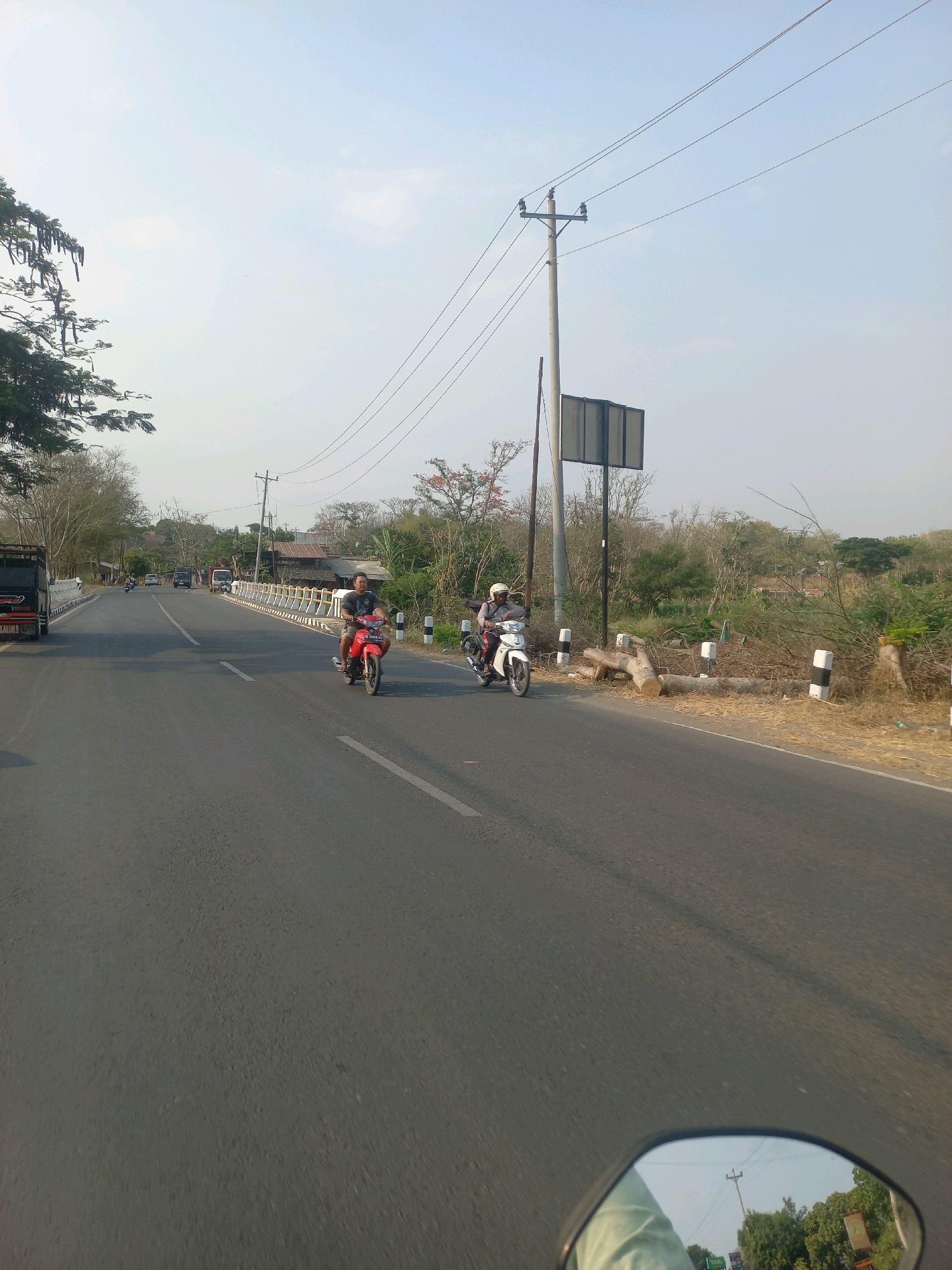 The image depicts a rural road scene in Baturetno, Indonesia, likely during daylight hours with a clear sky. The perspective is from a moving vehicle, possibly a motorcycle, as indicated by the reflection in the rearview mirror in the foreground. The road itself is paved with asphalt and features a white lane marker.
On the road, two motorcycles are visible. The first, closer to the viewer and to the right of the lane, is red and ridden by a man wearing a dark t-shirt and shorts. The second motorcycle is white and is positioned further ahead and to the left of the red motorcycle, also with a rider. Both motorcycles appear to be in motion.
The sides of the road are lined with vegetation, including trees and dry grass. There are several utility poles with overhead power lines crossing the scene. A large, rectangular billboard is mounted on one of these poles. Along the right side of the road, a low barrier with alternating black and white posts is present, separating the road from a dry, grassy embankment. Several large logs are scattered on this embankment. In the distance, buildings with tiled roofs suggest a settlement. A red truck is parked on the left side of the road further down. The overall impression is of a well-traveled but rural road.