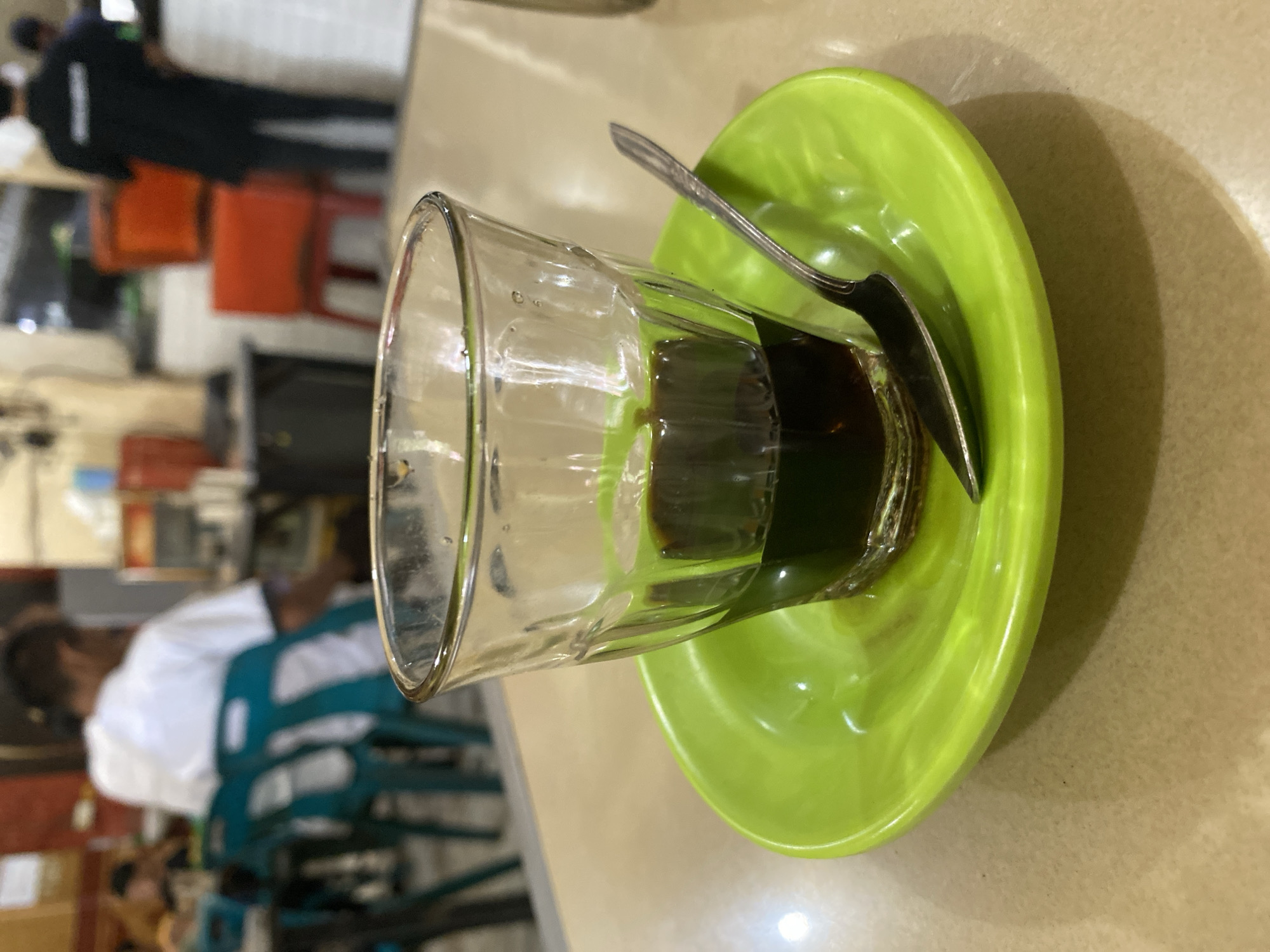 The image shows a glass of dark liquid, likely coffee, on a bright green saucer. A silver spoon rests on the saucer next to the glass. The scene appears to be in a casual eatery or cafe, with blurred figures in the background. In the middle ground, a person wearing a white shirt sits on a teal chair. In the background, a person in a black uniform stands behind a counter. The environment suggests a public space within Lhokseumawe, Indonesia, possibly a local coffee shop or food stall given the presence of simple furniture and the style of the drinkware. The lighting is artificial and appears to be from overhead fixtures.