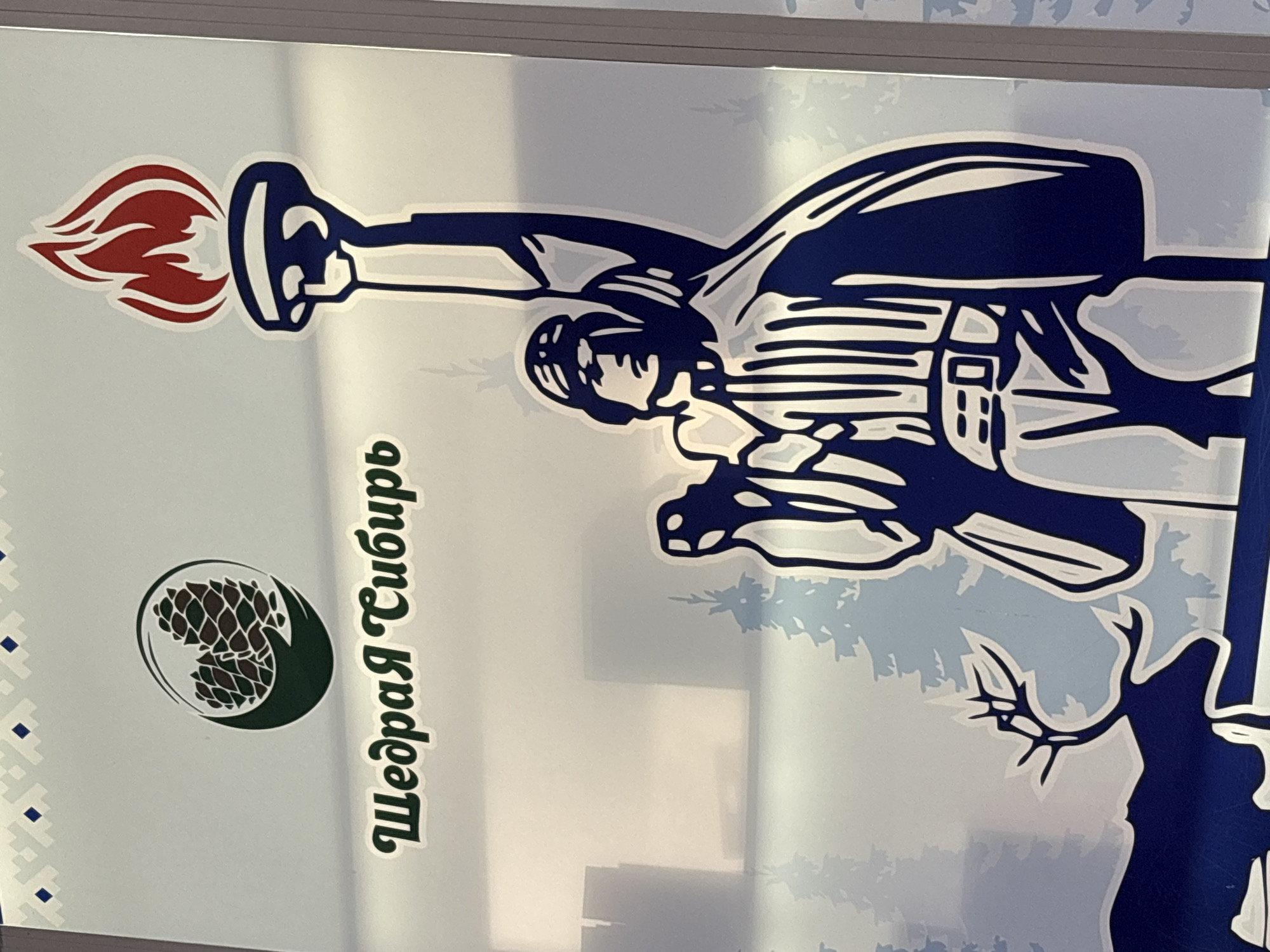 The image shows a sign for "Щедрая Сибирь" (Generous Siberia) in Nizhnevartovsk, Russia. The sign features a logo depicting a person with a torch and other elements, including what appears to be a pinecone or similar plant-based item and a graphic that suggests elements from a Siberian landscape. The person with the torch appears to be in a shooting pose, possibly with a rifle or similar long gun. The text "Щедрая Сибирь" is vertically aligned and written in a stylized font. The entire sign is framed.