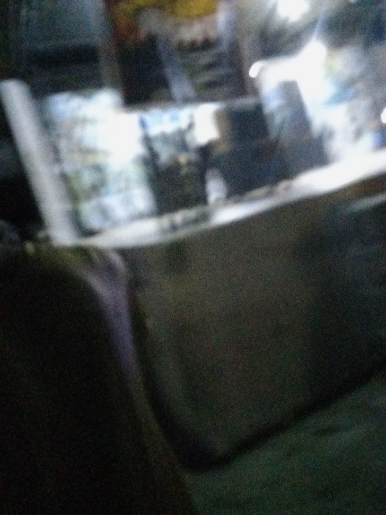 The image is blurry and taken at night. It appears to show a market stall or a small shop in Ado, Nigeria. There are several items on display, possibly food or household goods. A person is standing behind the stall. The surrounding area is dimly lit.