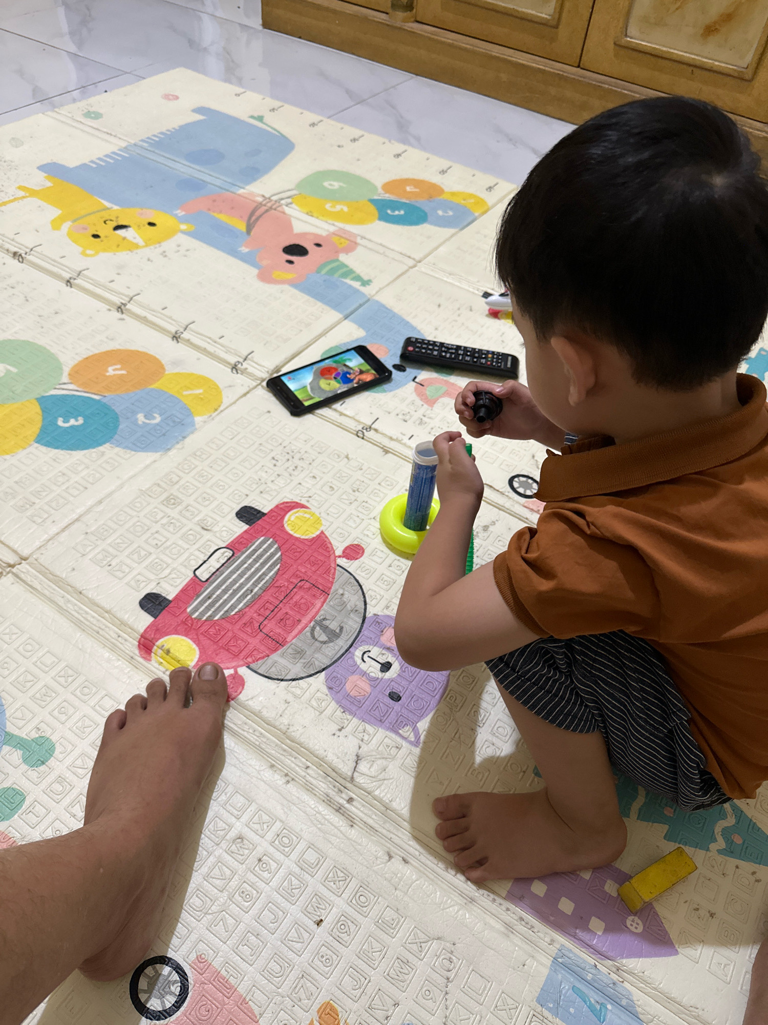 In Saruni, Indonesia, a young boy sits on a colorful play mat, engrossed in a smartphone. He wears an orange polo shirt and striped shorts, his dark hair neatly cut. A remote control lies nearby, alongside a small toy. The play mat depicts various cartoon animals and colorful circles with numbers. A man's foot is visible in the foreground, suggesting the presence of an adult. The scene appears to be an indoor setting, possibly a living room or playroom, with wooden furniture in the background.