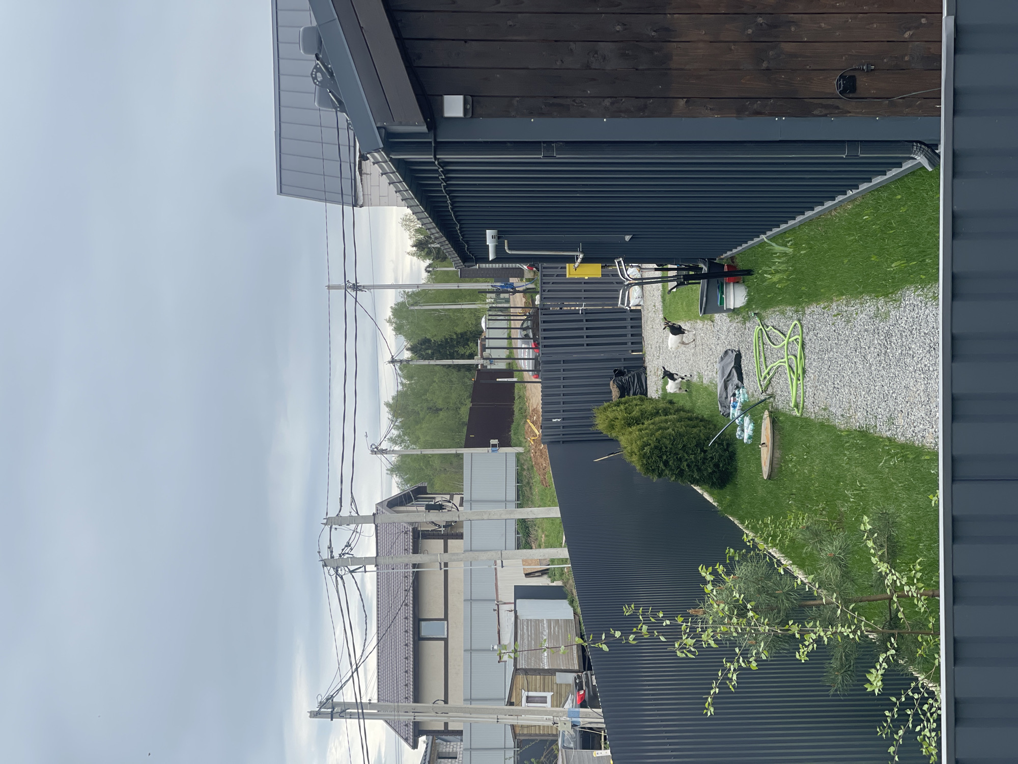 The image depicts a residential scene in Хлябово, Russia. The perspective is from an elevated position, looking across adjacent properties. In the foreground, a corrugated metal fence and the corner of a building with dark wood siding are visible. Between the fences, there is a narrow strip of land with gravel, grass, and various objects, including a green garden hose, lawn care equipment, and what appear to be plastic bottles and a trash bag. Two dogs, black and white, are present in the gravel area. Beyond the fences, there are more residential buildings, a gravel area, and an area of trees. Power lines stretch across the sky, supported by concrete poles. The sky is overcast and gray.