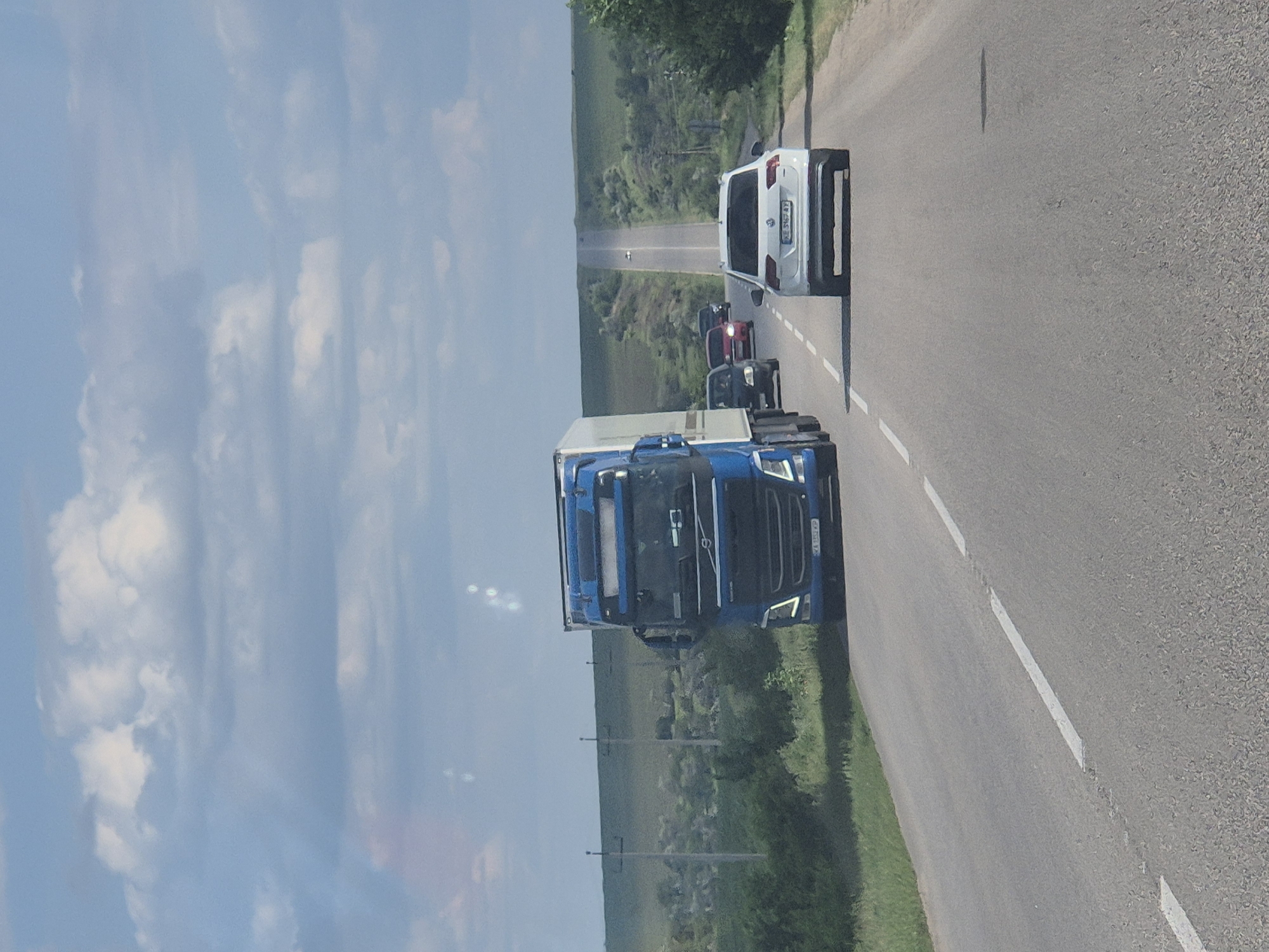 A blue Volvo semi-truck with a white trailer is traveling on a two-lane asphalt road, heading away from the camera. The truck is in the left lane. Ahead of the truck, in the right lane, is a white Volkswagen Tiguan. Further ahead in the distance, several other vehicles are visible, including a red car. The road is bordered by green grass and trees on both sides. In the background, rolling hills are covered with vegetation. The sky is blue with scattered white clouds. Electrical poles are visible along the side of the road. The scene appears to be a rural highway.