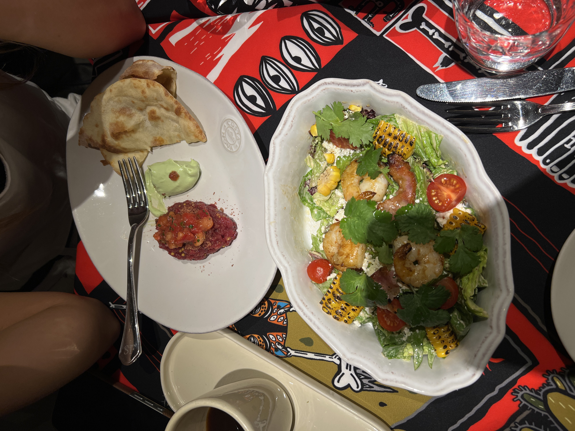 A high-angle shot shows two plates of food and a glass of water on a table covered with a patterned tablecloth.
On the left, a white plate holds a stack of pita bread, a dollop of green sauce, and a small mound of what appears to be raw meat. A fork rests on the plate.
To the right, a white bowl contains a salad with shrimp, tomatoes, grilled corn, lettuce, bacon and cilantro. A knife and fork are placed next to the bowl.
In the lower left, a person's arm and shoulder are visible. Below the plate on the left, there is a sauce dish and possibly a cup.
The patterned tablecloth features a mix of graphic designs in red, black, and white.
Based on other visual cues, the images seem to be from a restaurant in Moscow, Russia. This conclusion is drawn from the embossed 'Costa' logo on the left plate, which matches the style of dishes used in restaurants in the city, and based on the general food presentation.