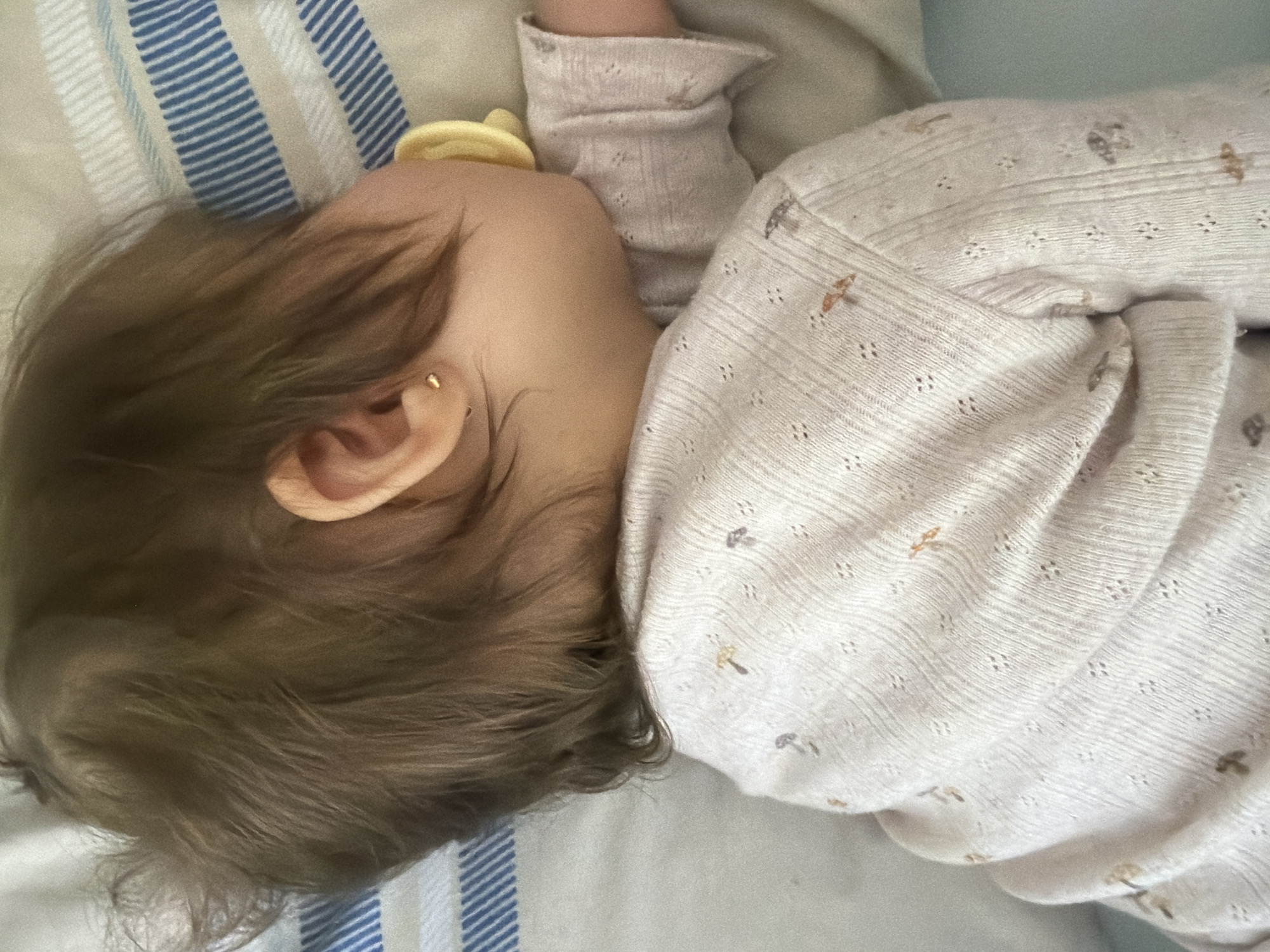A close-up, top-down image captures a baby or young child sleeping peacefully on their side. The focus is on the child's head, with their face turned away, revealing their ear and fine, light brown hair. A small, golden stud earring is visible in the earlobe. Part of a yellow pacifier can be seen near the child's mouth.
The child is dressed in a pale-colored, likely cream or light pink, long-sleeved top or sleepsuit made of a soft, textured fabric. The garment features tiny patterned holes and small, scattered motifs in muted brown and grey tones, resembling leaves or insects.
The child rests on bedding consisting of a light-colored pillow or sheet with blue and white stripes, and another plain light-toned sheet or blanket. The scene conveys a sense of calm and rest, consistent with a child sleeping indoors, likely in a bedroom. The lighting is soft and diffused. No specific details within the image indicate the provided location of Bearpark, United Kingdom. There is no visible text.