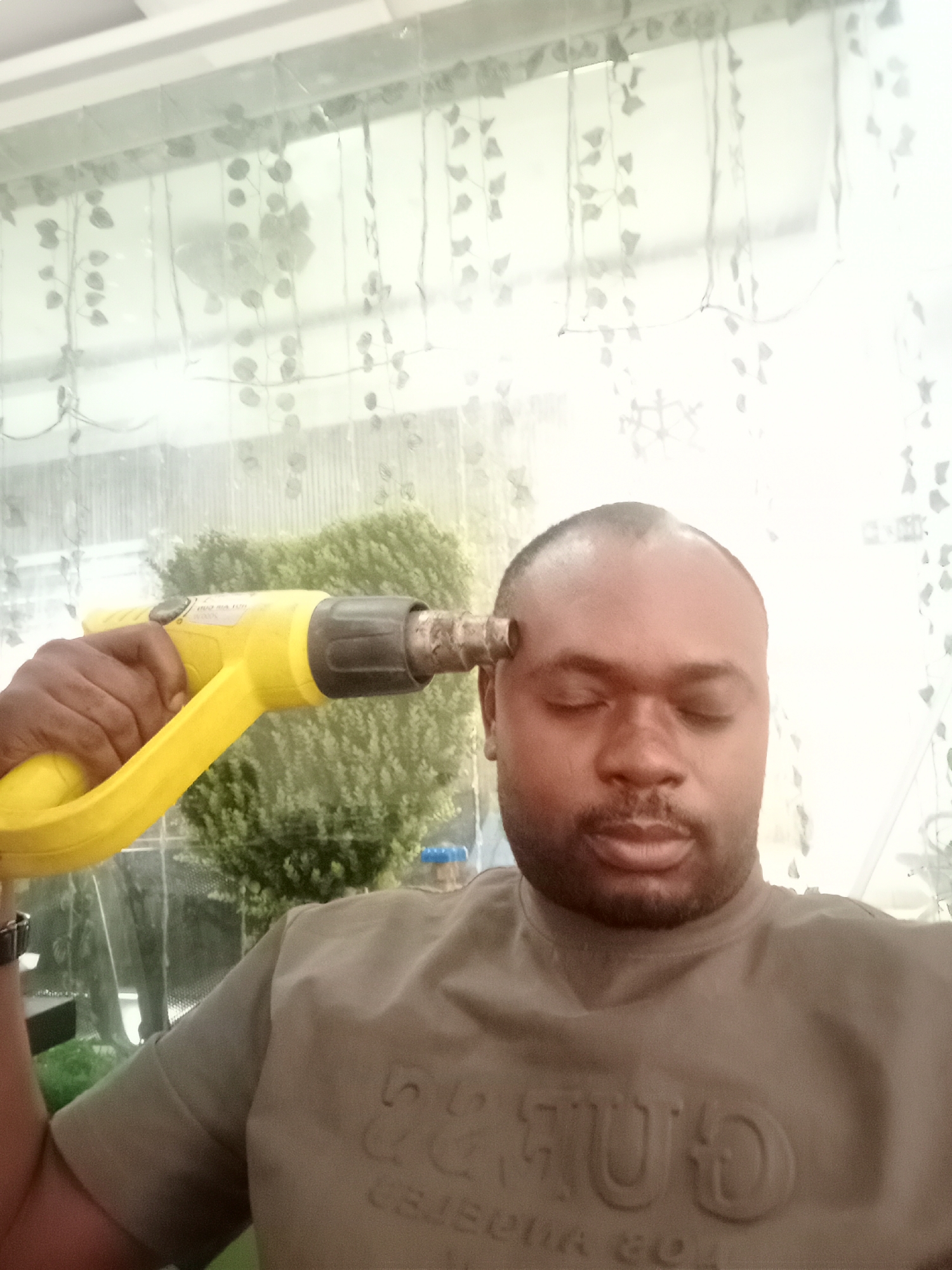 An indoor, vertical shot captures a Black man holding a bright yellow heat gun to his right temple, with his eyes closed. The man has a dark complexion, short hair (with some thinning on top), and a mustache. He is wearing a light brown, short-sleeved top with embossed letters across the chest; visible parts suggest words like "TUESDAY" and possibly "CALIFORNIA" or "COLLEGE."
The heat gun, primarily yellow with black components, has partially legible text on its side, including elements of the brand "KÄRCHER," "TYPE," "max. 60°C," and "IPX4." The nozzle of the device is pressed against his head.
The background consists of sheer white curtains featuring a delicate pattern of light-colored leaves or vines, which appear to be hanging in front of a window or glass partition. Through the translucent curtains, lush green foliage is visible, suggesting either an outdoor garden or significant indoor plants. The scene is well-lit, indicating daytime conditions.
The man's closed eyes and the symbolic gesture of holding a heat gun to his head could suggest metaphorical stress, exhaustion, or perhaps a humorous and ironic representation of feeling "overheated" or burnt out. The setting appears to be an interior space, possibly a home or business in Gwarinpa, Nigeria.