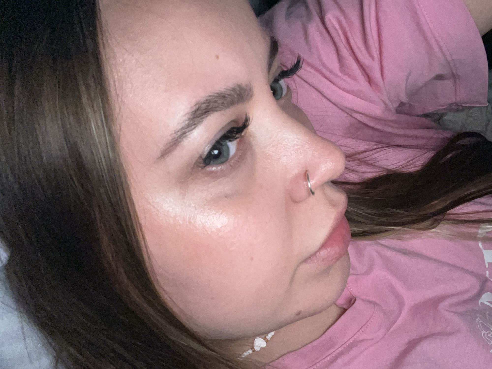 A close-up photograph captures a woman's face and upper torso. She is turned slightly to the right, with her gaze directed upwards and to the right. Her hair is dark brown with lighter brown highlights. She has fair skin with rosy cheeks, and a silver nose ring. She is wearing false eyelashes. A white beaded necklace is visible around her neck. She is wearing a pink t-shirt. The setting is likely indoors. The location in Novopokrovskaya, Russia is unspecified.