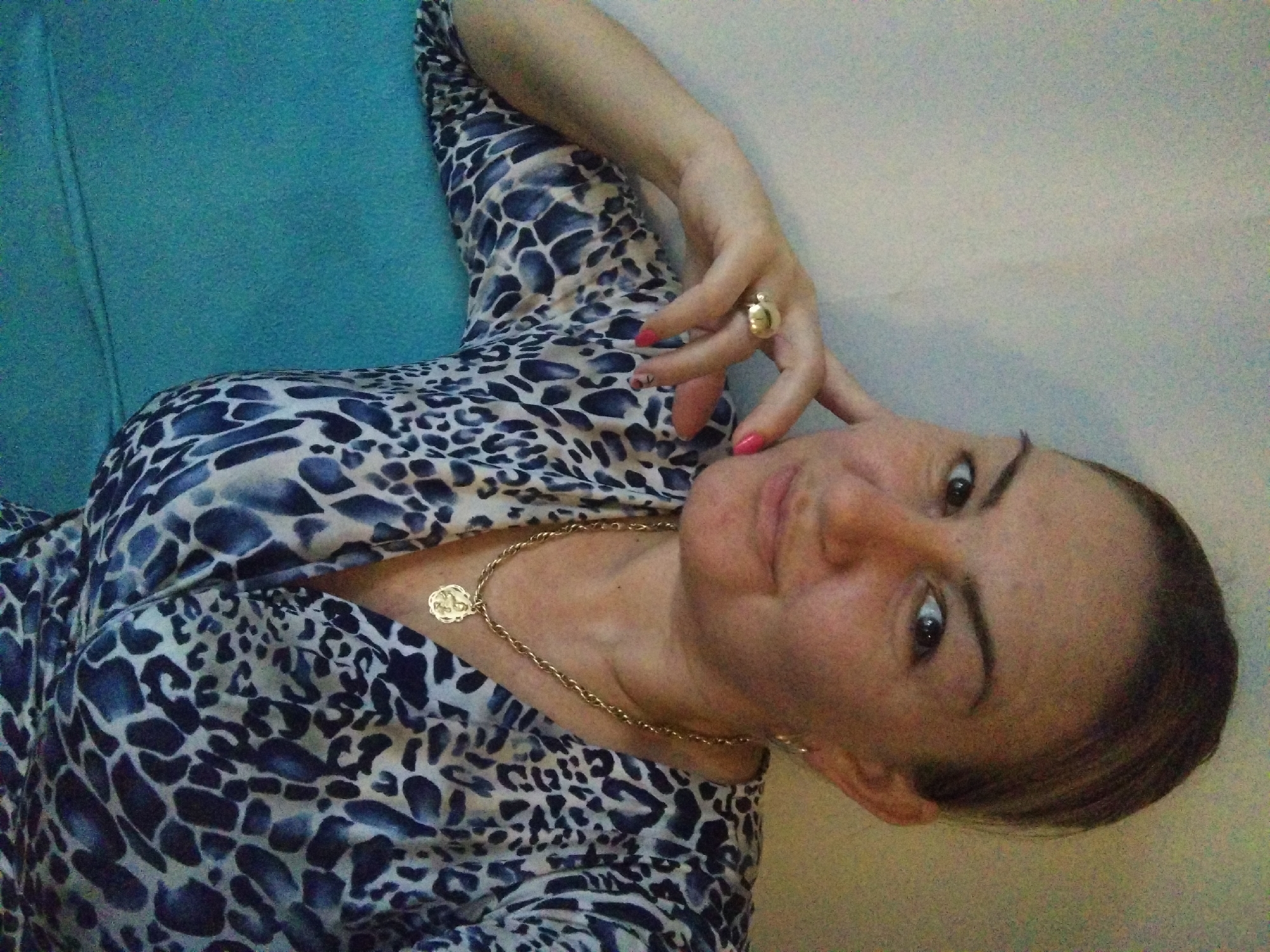The image is a close-up shot of a woman lying down, possibly on a blue surface, against a light-colored wall. She is wearing a patterned dress, a gold necklace with a round pendant, and a large gold ring. Her hand is near her face, and she is looking at the camera with a slight smile. The location is not discernible from the image, and it is not possible to determine if the photo was taken in Stara Zagora, Bulgaria.