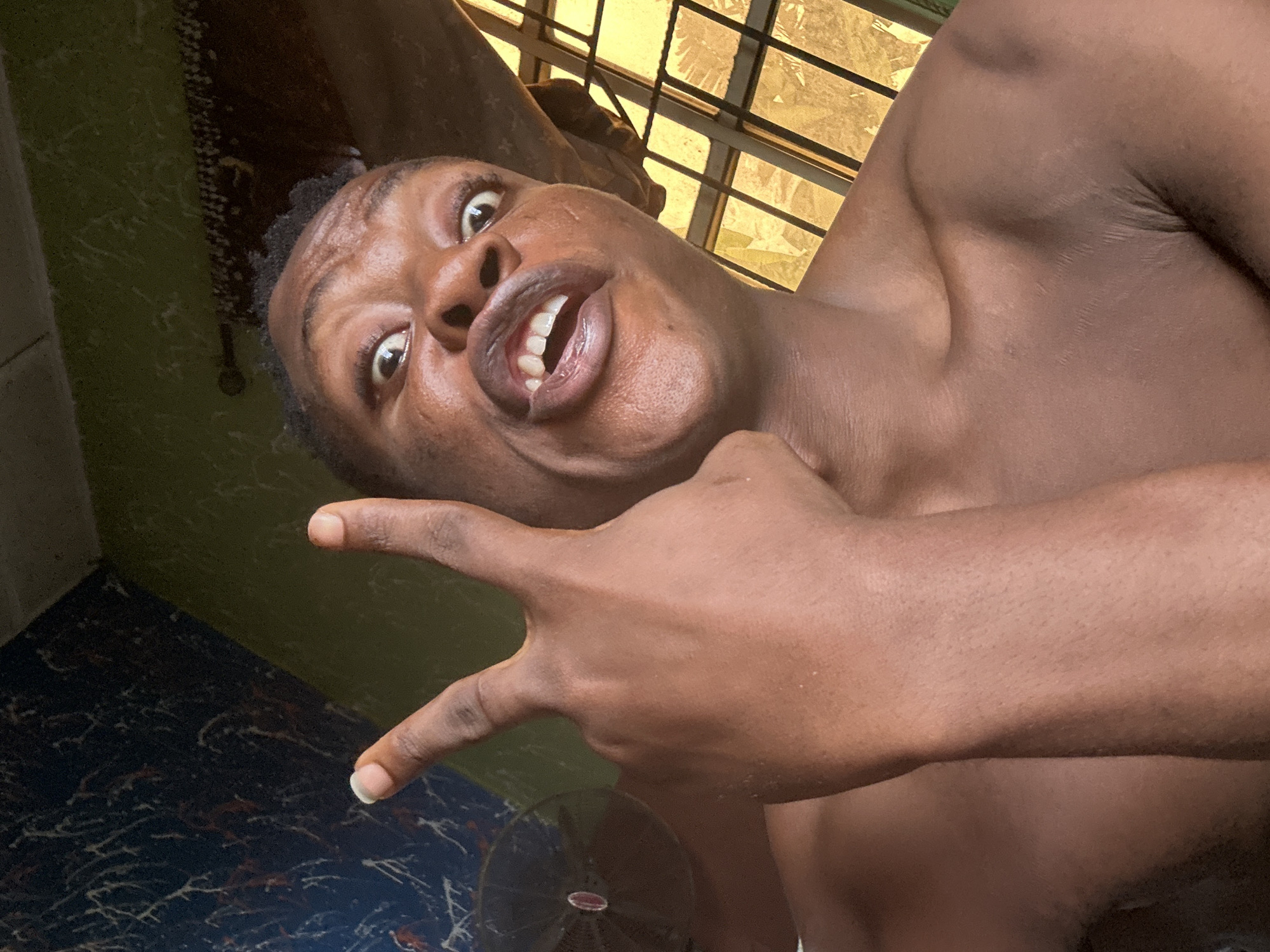 The image is a close-up, low-angle selfie of a young Black person. The subject has dark skin, short, curly dark hair, and is making a peace sign with their right hand in front of their face. They have a wide-mouthed expression, showing teeth, and their eyes are wide open, looking directly at the camera. The background appears to be an indoor space with patterned walls, one section a dark blue with white and red streaks, and another section a muted green. To the right of the subject, there is a window with a yellow tint and horizontal bars. A circular fan with a red center is visible to the left. The overall lighting suggests it is daytime, possibly near a window. The location is specified as Ikorodu, Nigeria.
