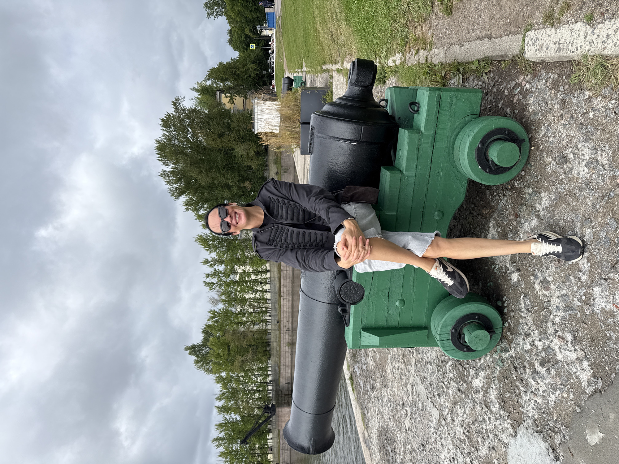 A person is sitting on the carriage of a large, black cannon. The cannon is positioned on a concrete surface next to a body of water. The cannon carriage is painted bright green and has two large, wooden wheels on each side. The person is wearing sunglasses, a black jacket with decorative braiding, light-colored shorts, and athletic shoes. They are smiling and looking towards the camera. In the background, there are trees, buildings, and a cloudy sky. The location is identified as Kronstadt, Russia.