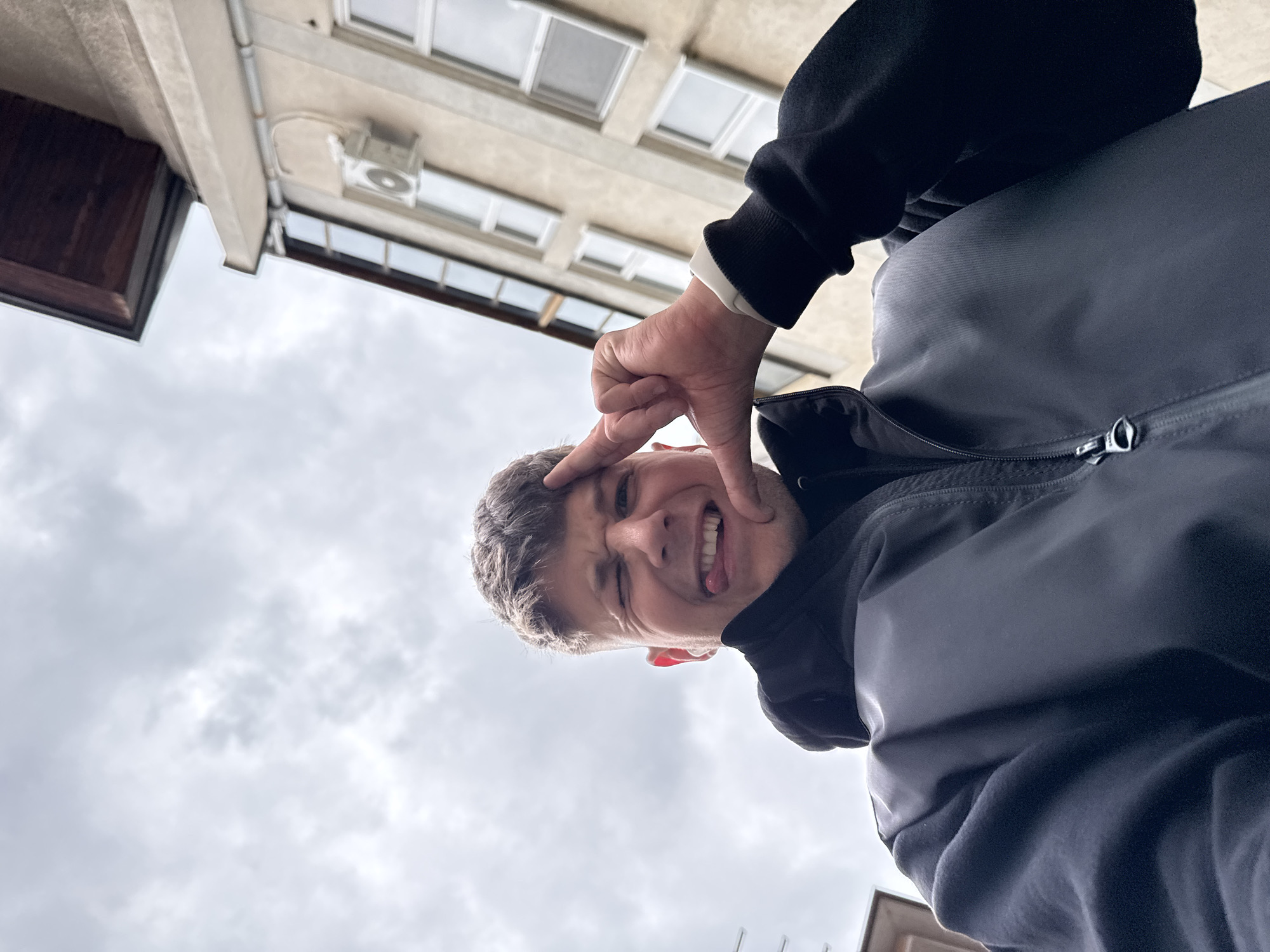 A low-angle, rotated outdoor photograph captures a young man making a playful expression, set against an overcast sky and the exterior of a building in Plovdiv, Bulgaria. The image is rotated counter-clockwise, showing the subject and background on their sides.
The primary subject is a young man, visible from the chest up, positioned in the foreground and center-right. He is looking towards the camera, winking with his left eye, sticking out his tongue slightly, and smiling broadly. His right hand is raised, with his index and thumb extended to form an "L" shape or a stylized "loser" sign, pointing towards his right temple. He is wearing a dark-colored jacket, possibly black or navy, over a dark long-sleeved shirt, with a white cuff visible on his right wrist. His hair is short and brown.
The background features an overcast sky, filling the lower portion of the rotated image. Above the sky, partially obscured, is the underside of a building's overhang or balcony, along with a beige wall featuring several rectangular windows. A white air conditioning unit or similar fixture is attached to the wall. A thin, light-colored pipe or conduit runs horizontally along the building. On the far left, a dark brown, possibly wooden, architectural element is visible, suggesting a decorative trim or rafter.
The overall mood is lighthearted and cheerful, conveyed by the man's expression and gesture. The weather appears cloudy, indicating daytime without direct sunlight. There is no visible text in the image.