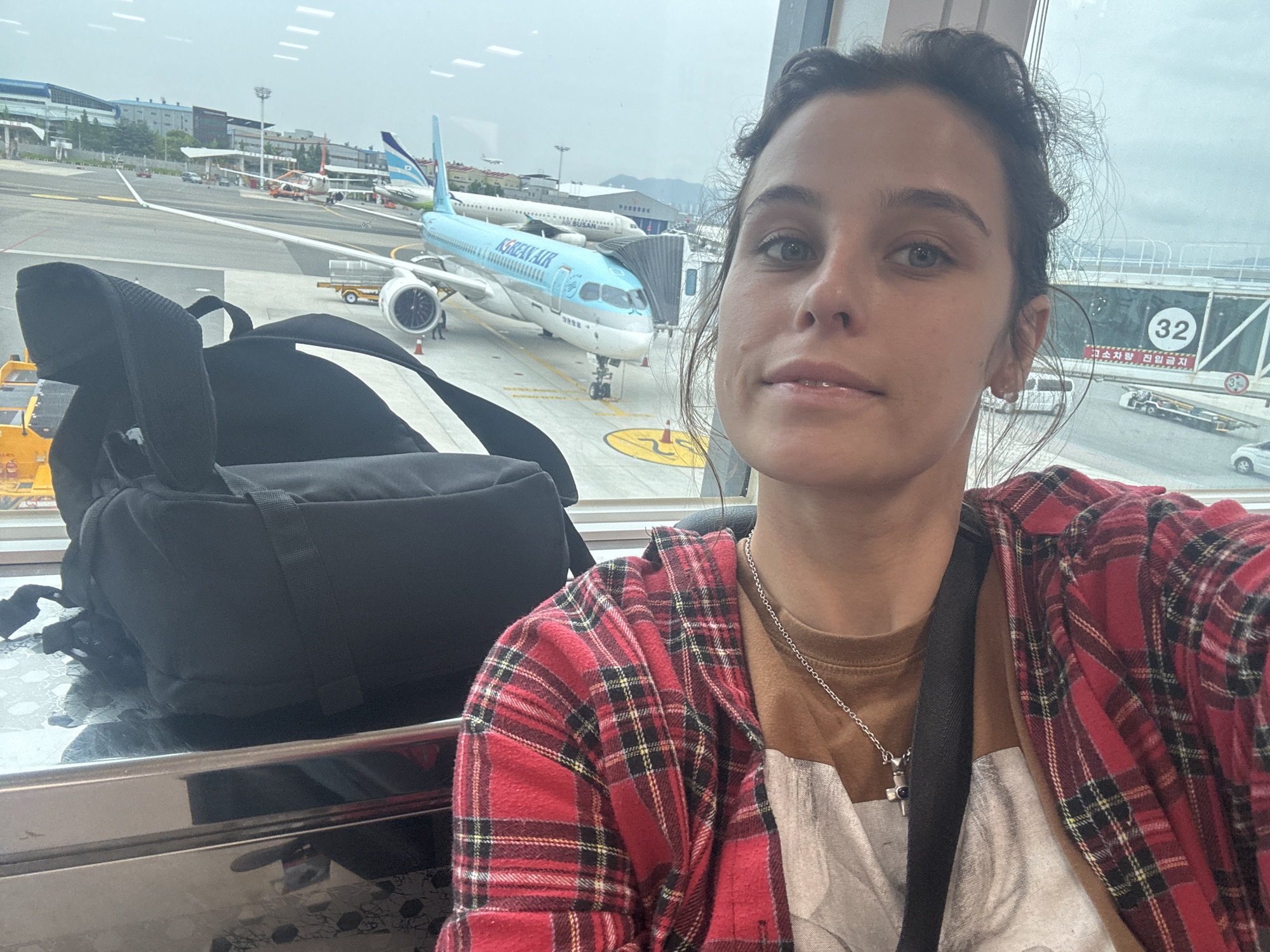 A young woman with brown hair is taking a selfie inside an airport terminal. She is wearing a brown t-shirt and a red and black plaid shirt over it, along with a silver necklace. A black backpack is visible to her left, and a shiny, patterned surface is below. Through the window behind her, a blue Korean Air airplane is parked at a gate. The airport environment with the airplane and tarmac is visible. Signs in Korean and English are present. The scene appears to be at Gimhae International Airport, serving Busan, South Korea.
