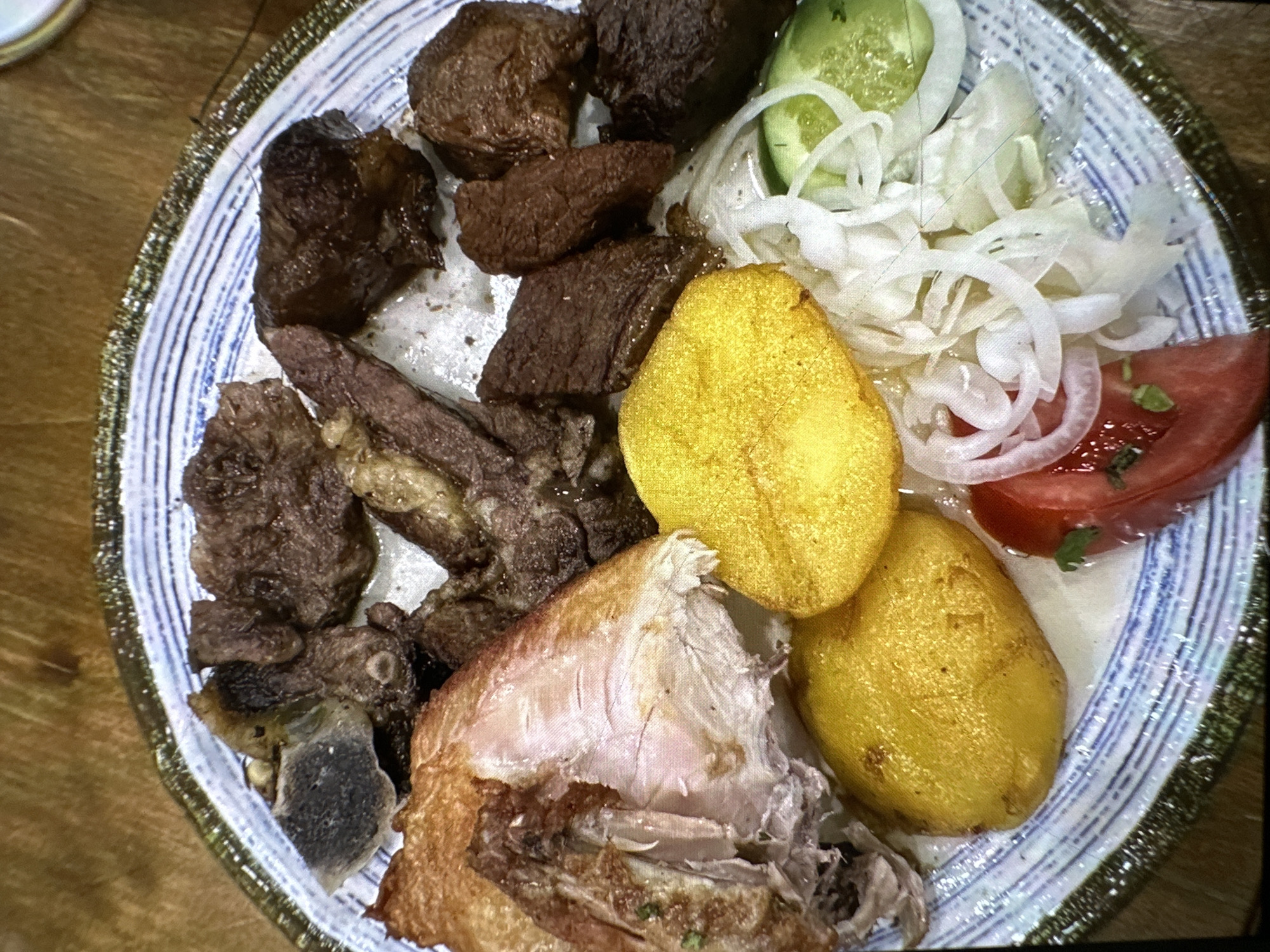 A close-up overhead shot shows a round, blue and white patterned plate filled with food. The plate is placed on a wooden surface. The food consists of several pieces of dark, roasted meat, some with bone fragments, a large piece of roasted chicken with crispy skin, two golden-brown roasted potato halves, a slice of tomato with green garnish, thinly sliced white onions, and a few slices of cucumber. The lighting is warm, creating shadows and highlighting the textures of the food. No people or activities are visible in the image. The location is specified as Кызыл-Булак, Kyrgyzstan.