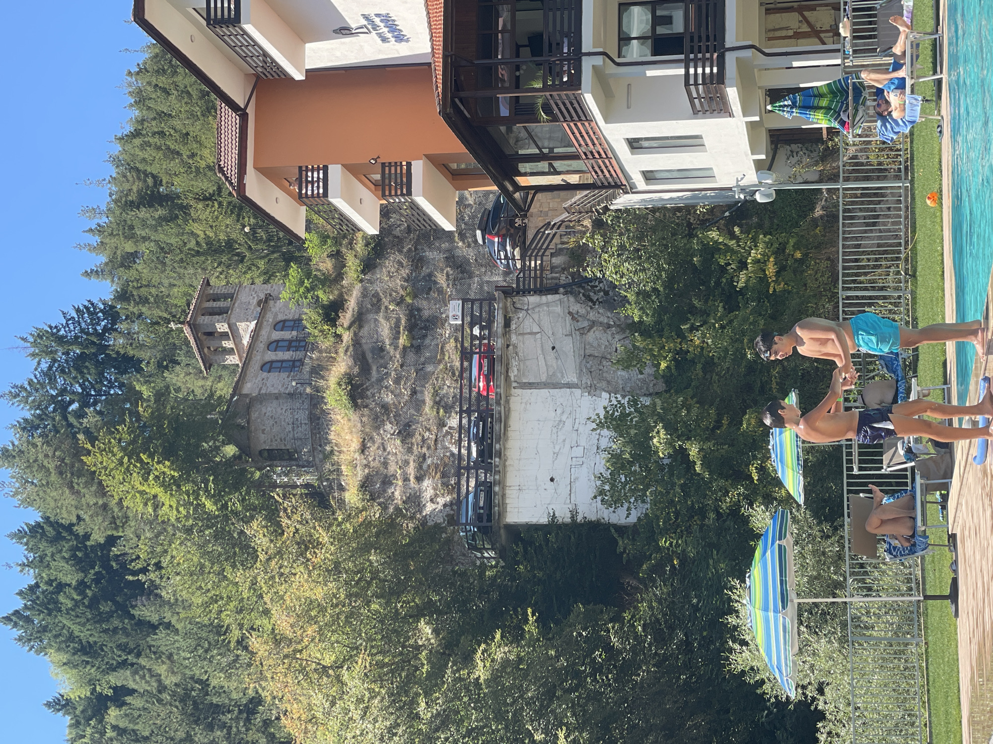 The image shows a view of the Rila Monastery in Banite, Bulgaria, nestled on a hillside covered with lush green trees. A part of the monastery, featuring stone walls and arched windows, is visible at the top of the frame. In the foreground, there is a swimming pool area with two young boys standing near the edge. One boy is wearing blue swimming trunks, and the other is wearing dark blue swim shorts. There are lounge chairs, umbrellas, and people relaxing by the pool. To the right of the pool, a multi-story hotel building with balconies and orange and white facades is visible. A road with parked cars is seen on a raised platform on the hillside, above the pool area. The overall scene depicts a sunny day with a blend of historical architecture and modern leisure facilities.