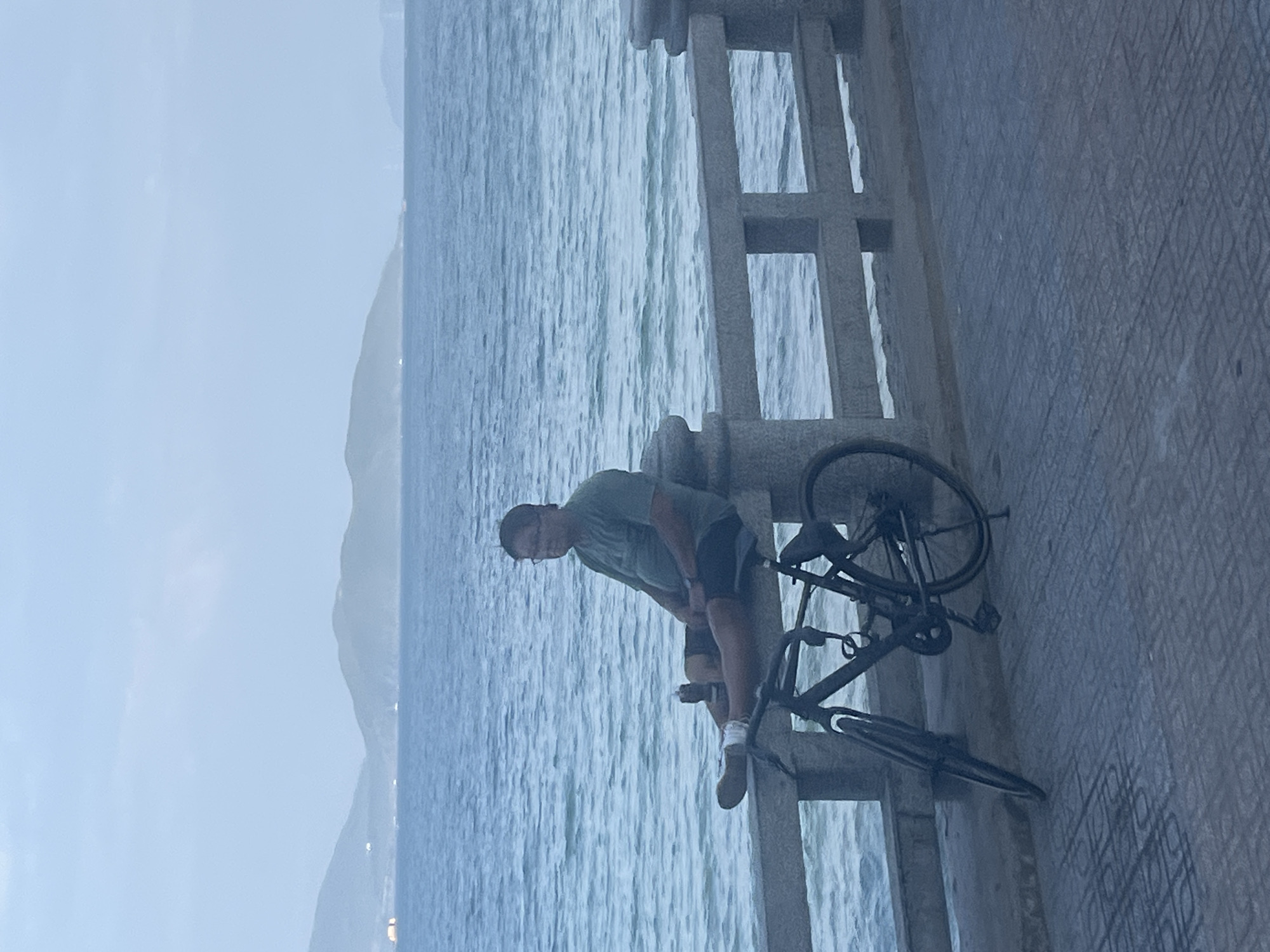 A middle-aged man wearing glasses, a green t-shirt, and dark shorts sits on a stone railing overlooking the sea. He has a bottle in his hand and is resting his legs on the railing. A bicycle is parked to his left, leaning against the railing. The sea is calm and blue, with hills visible in the distance under a hazy sky. The scene appears to be a public promenade in Nha Trang, Vietnam, likely along Tran Phu Beach, given the coastal setting and the presence of a pedestrian walkway with railings and the sea.