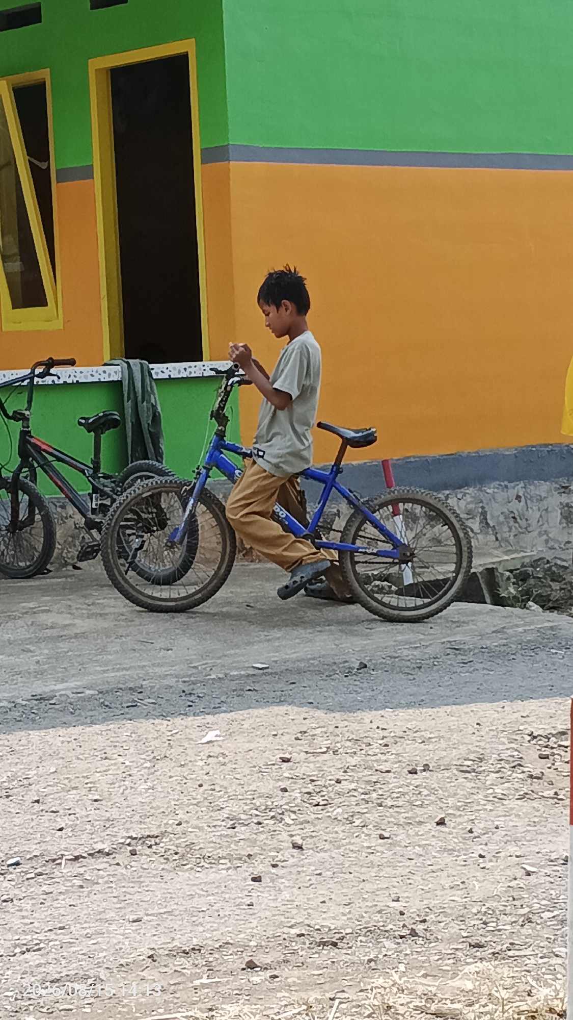 A young boy in a light brown t-shirt and tan pants sits on a blue BMX bicycle in front of a building in Desa Rawajaya, Indonesia. He appears to be engaged with his phone. To his left, another black bicycle is parked. The building behind them is painted with horizontal bands of bright green, gray, and orange. A dark doorway and a window with yellow trim are visible on the left side of the building. The ground in front of the building is a mixture of gravel and dirt, with shadows indicating a sunny day. The overall scene suggests a casual, outdoor setting.