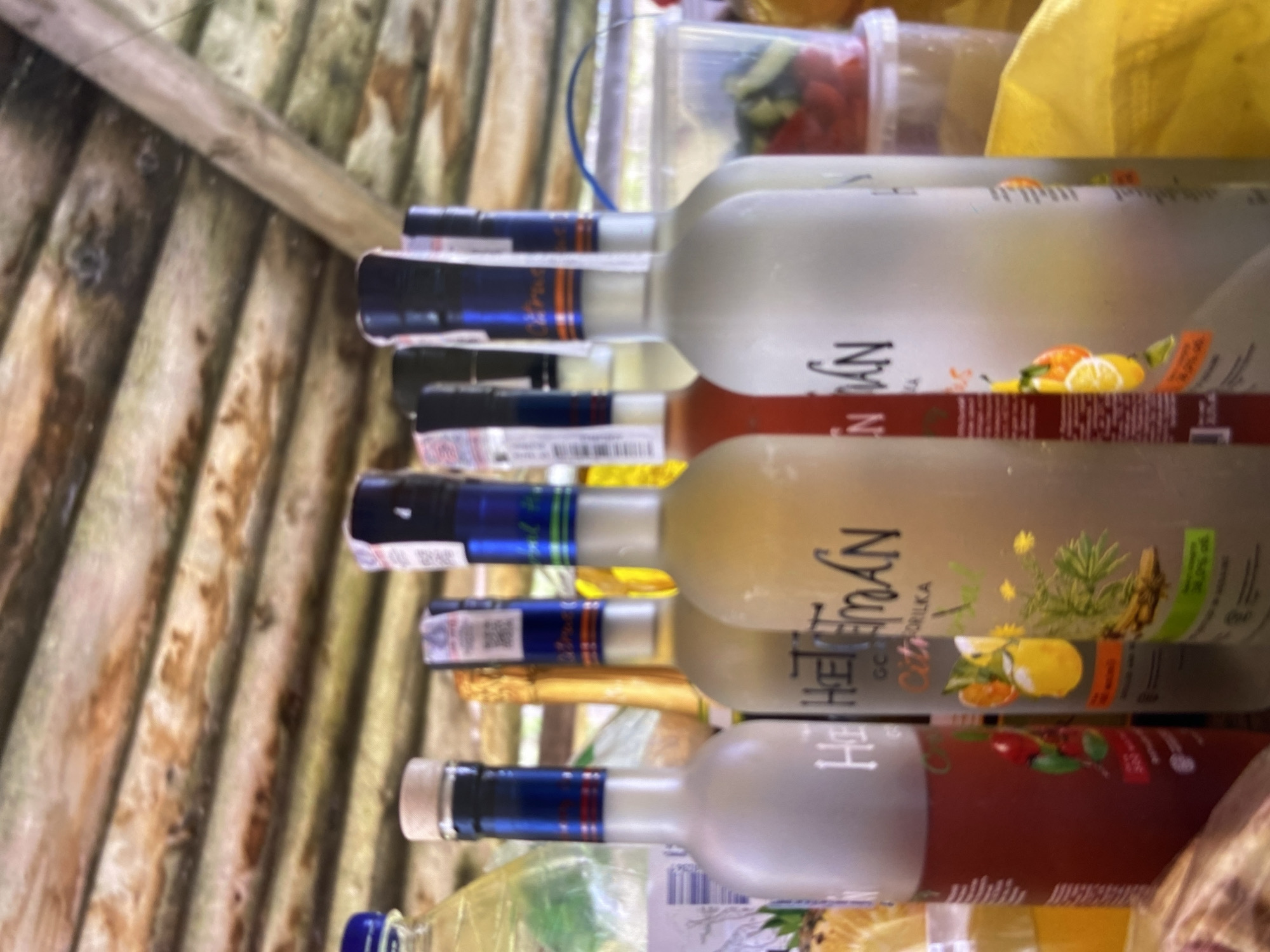 The image shows several bottles of alcohol arranged on a table under a wooden roof. The bottles are of various shapes and colors, some with visible labels. In the background, there is a clear plastic container filled with what appears to be chopped vegetables, possibly for a salad or garnish. The overall setting suggests an outdoor gathering or picnic.
