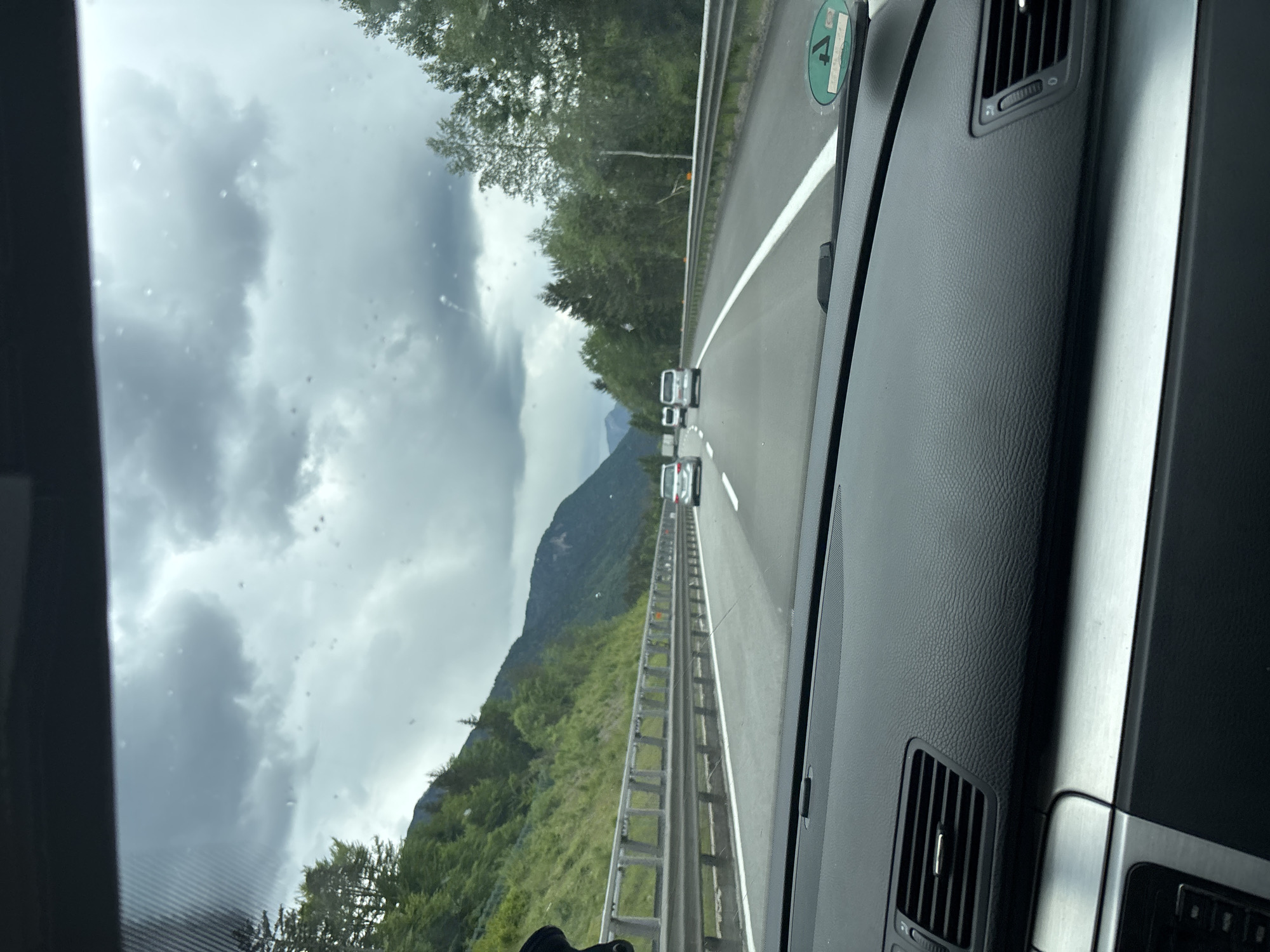 The image shows a view from inside a car driving on a highway in Malborghetto-Valbruna / Naborjet-Ovčja Vas / Malborgeth-Wolfsbach / Malborghet e Valbrune, Italy. The car is traveling on a multi-lane road with a white dividing line and metal guardrails. Other vehicles are visible ahead. In the background, there are mountains and trees under a cloudy sky. The dashboard of the car is visible on the right side of the frame. A green sticker is visible on the windshield, likely a road tax or environmental permit for Austria.