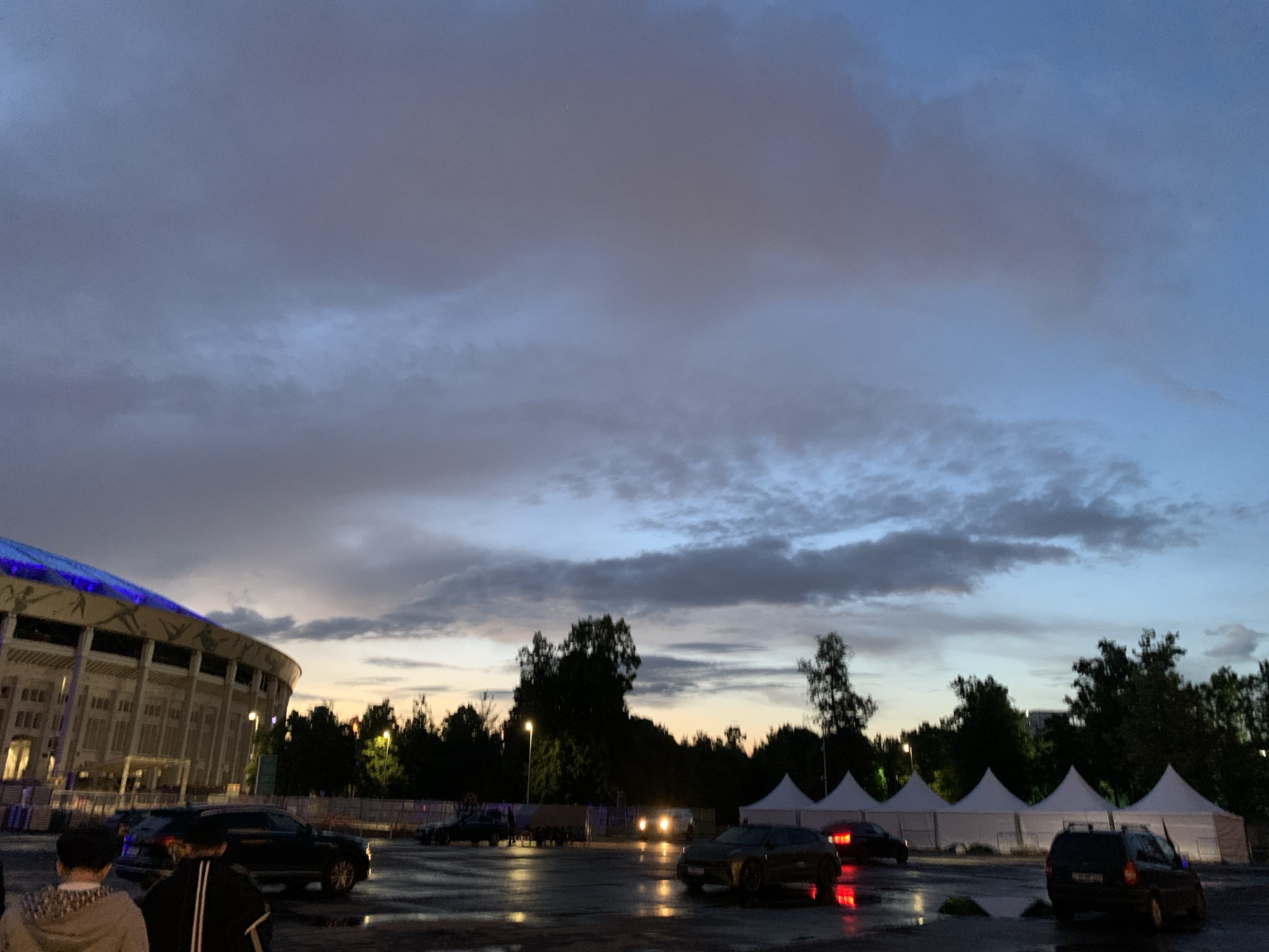 The image depicts an outdoor scene at dusk, possibly after rain, given the wet ground reflecting light. A large stadium with a blue-lit structure is on the left side of the frame. Trees are in the midground. Several cars are scattered across a parking lot. The background features white tents. Two individuals are partially visible in the foreground on the left. The location is a parking lot outside of the stadium. The sky is mostly cloudy. The location is in the city of Unknown, Unknown.