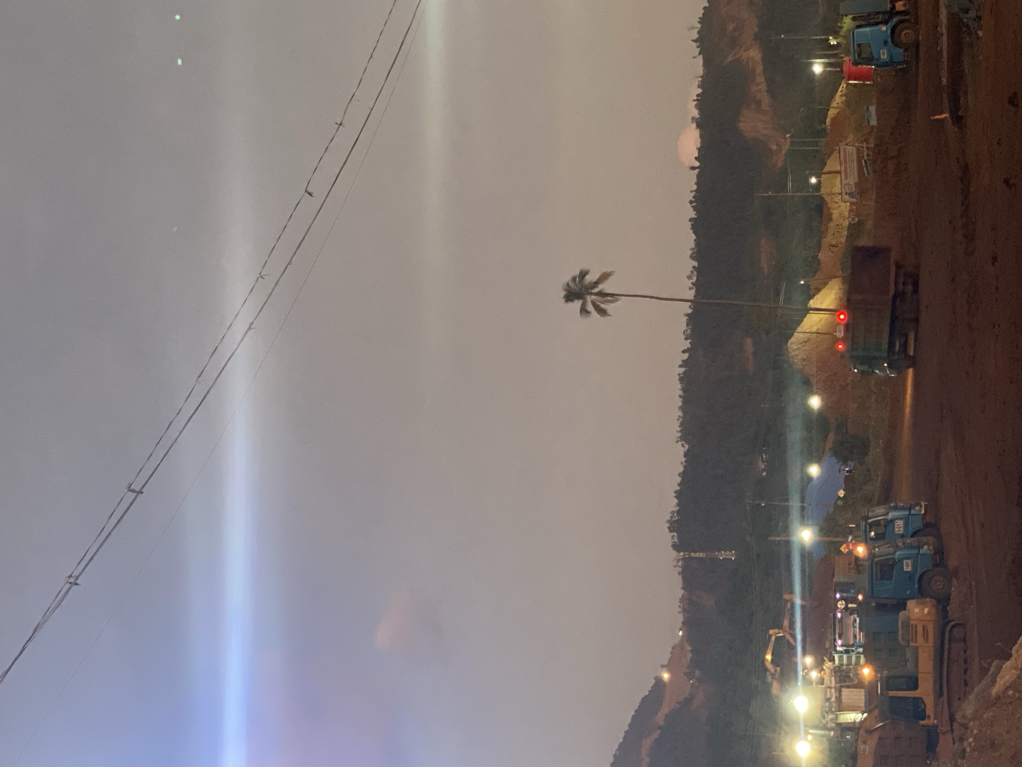 This is a nighttime or dusk photograph taken at a construction or mining site in an unspecified location in Indonesia. The foreground is dominated by dark brown earth and a yellow excavator parked on the left. To its right, several blue dump trucks are parked, with their headlights and hazard lights illuminated. One truck in the middle has its brake lights illuminated red. In the middle ground, there are large piles of dirt or sand, some covered with blue tarps. Various construction lights are illuminating the scene, casting a hazy glow. In the background, a densely forested hillside rises, with some lights visible on its slopes, suggesting settlements or further construction. A solitary palm tree stands tall in the middle distance. Overhead, power lines are strung across the sky. The sky is a muted, hazy gray with a strong lens flare emanating from the left side, suggesting a bright light source just out of frame. The overall atmosphere is industrial and busy, despite the low light conditions.