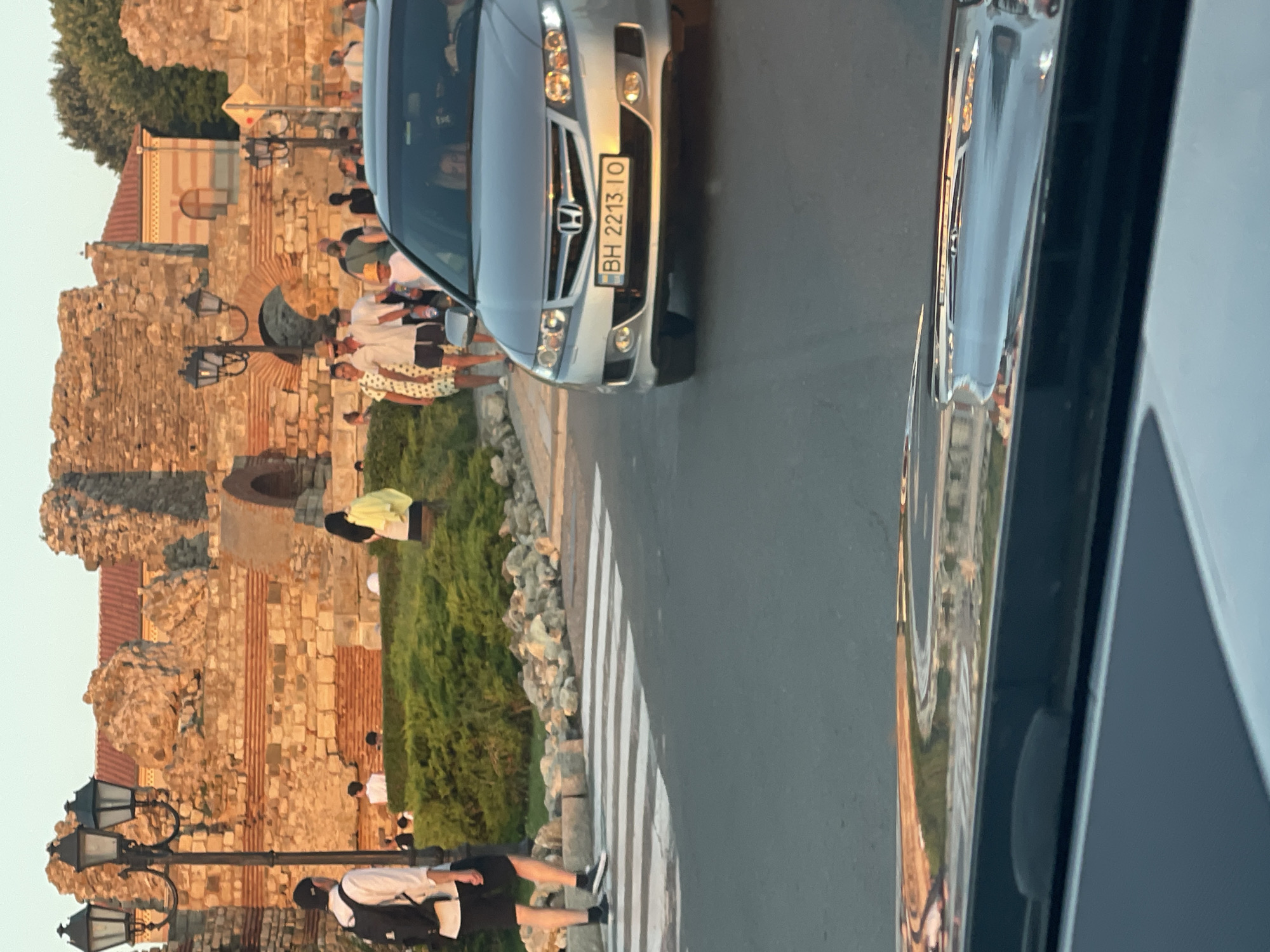 A silver Honda Accord car with Bulgarian license plate BH 2213 IO is traveling on a paved road. The car is in the foreground, right of center. In the background, to the left, is a pedestrian crossing. Beyond the crossing, a man wearing a white shirt and black shorts with a backpack is walking. Further back, a group of people are walking and standing near an ancient stone wall, some of which is covered by red-tiled roofs. The setting appears to be a historic area in Nesebar, Bulgaria, possibly near the old town, with characteristic lampposts and stone architecture. The lighting suggests it is late afternoon or early evening. The image is taken from inside another vehicle.