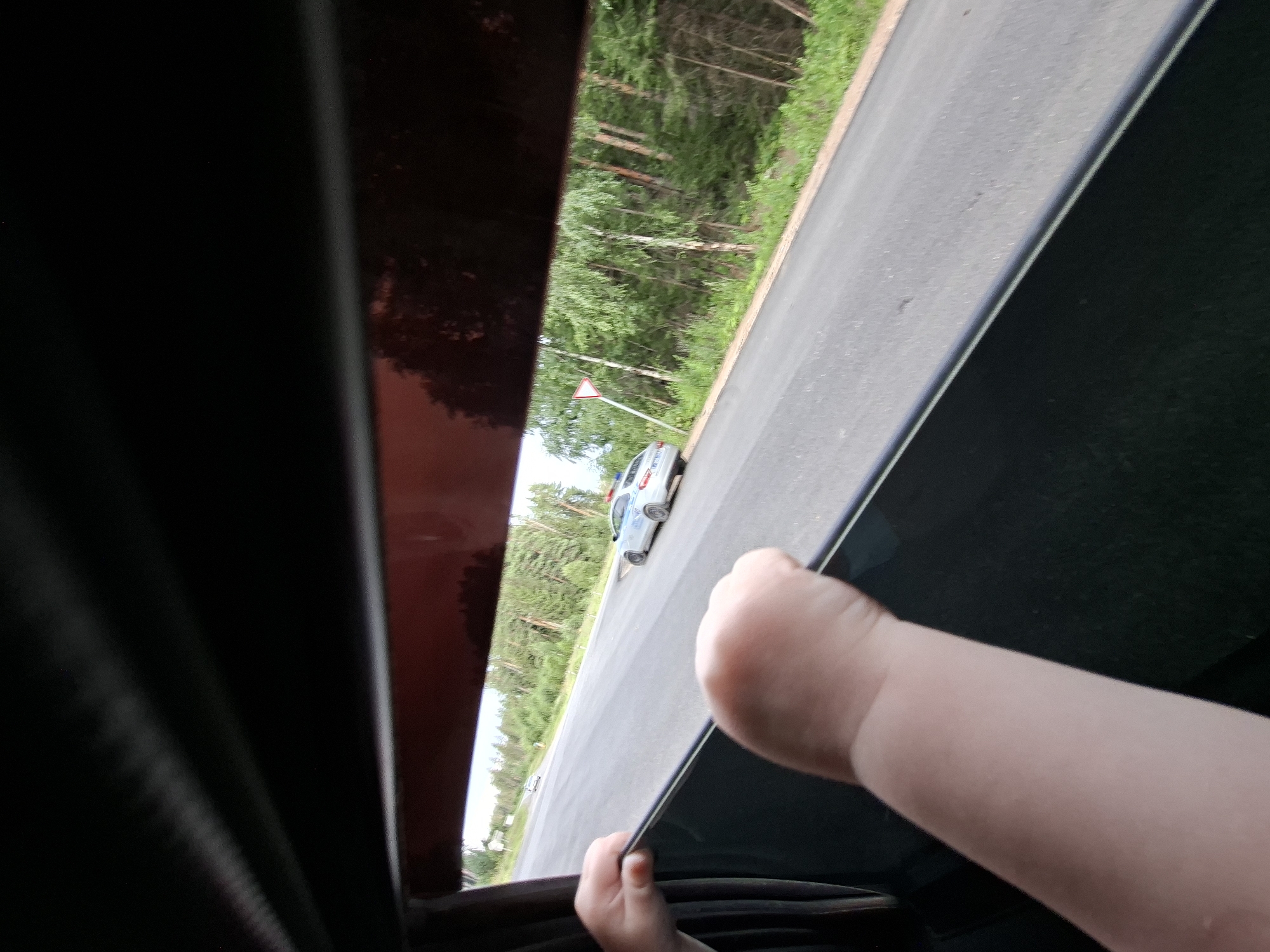 The image is taken from inside a vehicle, looking out of the window. A child's arm and hand are visible in the foreground, resting on the window frame. The window appears to have tinted glass or a sunshade. Outside the vehicle, a paved road is visible, bordered by green grass and trees. A white car, possibly a police vehicle with lights on top, is driving on the road in the middle ground. Further down the road, another white car is visible in the distance. A triangular yield sign is posted on the side of the road. The overall environment suggests a rural or suburban setting with dense forestation. There is no specific information to indicate the location is in the city of Votkinsk, Russia, or any other specific city.