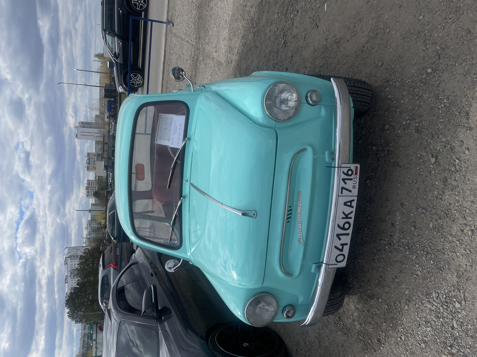 The image shows a light blue ZAZ-965 car with the license plate 0416KA 716, indicating it is registered in Russia. The car is parked on a gravel-covered area, suggesting an informal parking spot. Other vehicles are parked nearby, including a black SUV. Buildings are visible in the background, hinting at an urban setting. The sky is overcast with clouds. The sign on the windshield reads "ИЩУ ДЕВУШКУ НА КЛУБ", which suggests that the owner is looking for a girl to go to a club with. There is no readily available information to pinpoint the exact location in Naberezhnye Chelny.