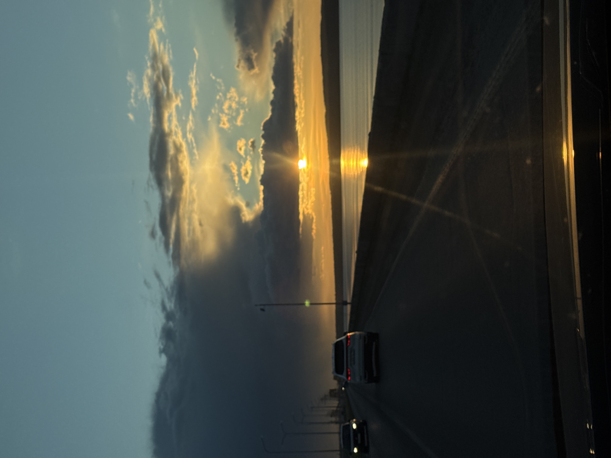 The image depicts a sunset scene over a body of water, likely the Kama River near Chaikovsky, Russia. The view is taken from inside a moving vehicle, as evidenced by the road, other vehicles, and possible window frame visible on the right side. The sky is split between dark, heavy storm clouds and areas illuminated by the setting sun. Light posts line the road. Reflections of the sun are visible on the water's surface. The overall atmosphere is dramatic due to the contrast between the storm and the sunset.