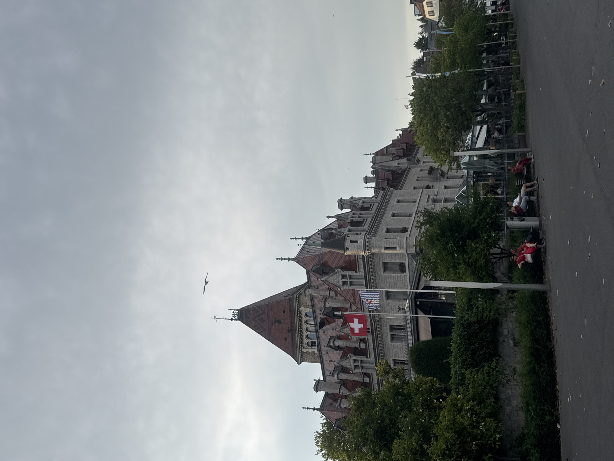 The image captures a view of the Beau-Rivage Palace in Lausanne, Switzerland, situated along the shores of Lake Geneva. The grand hotel, characterized by its intricate architectural style with mansard roofs, dormer windows, and ornate stonework, dominates the upper portion of the frame. A Swiss flag and another flag with blue and white stripes are prominently displayed on the hotel's facade.
In the foreground, a paved walkway runs alongside a lush green area populated by various trees and dense bushes. A stone wall separates this green space from the hotel grounds. Two people are seated on a bench within this green area, facing away from the camera and towards the hotel or lake. One individual wears a red top and another a white top. Further along the walkway, beyond the green area, a series of outdoor seating arrangements, likely for a cafe or restaurant, are visible under awnings.
The sky above is overcast with a pale, diffused light. A lone bird is captured in flight in the upper portion of the image, silhouetted against the sky. The overall scene suggests a calm, late afternoon or early evening atmosphere.