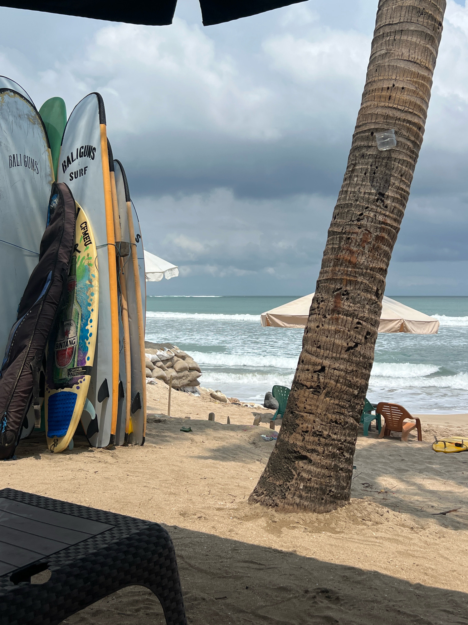 This image depicts a beach scene in Kuta, Indonesia. In the foreground, a dark woven table is visible. The middle ground features a row of surfboards leaning against a wall, some with brand names like "BALIGUNS" and "SURF". A surfboard with a design of a Bintang beer bottle is also present. To the right, a large palm tree trunk dominates the frame, with a plastic bottle attached to it. Beyond the palm tree, the ocean with waves is visible, along with a beach umbrella and several chairs. In the distance, sandbags line the shore, and the sky is overcast. The overall atmosphere is one of a relaxed, tropical beach environment.