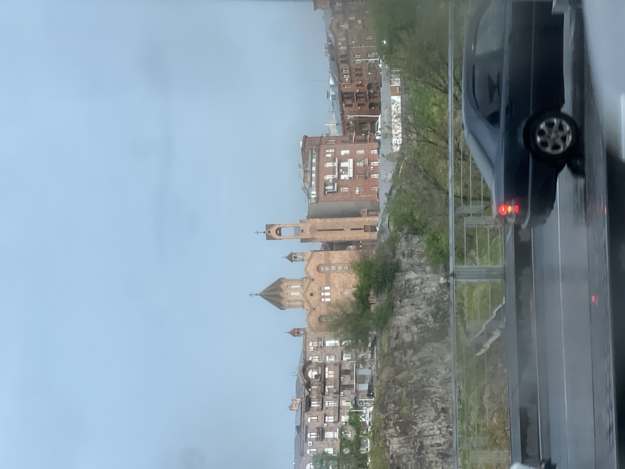 The image captures a view from a vehicle on a rainy day in Yerevan, Armenia, overlooking the architectural landscape.
Foreground: The right side of the frame shows the side of a dark-colored car with its wheel visible, suggesting the photo was taken from inside a moving vehicle. The wet pavement reflects the surrounding lights.
Midground: A stone cliff with some vegetation is visible. A church-like building with towers and crosses sits on top of the cliff, appearing to be part of a religious complex. Residential buildings are seen adjacent to the church.
Background: Further away, the cityscape of Yerevan is visible with multiple buildings of different heights and architectural styles. The sky is overcast, contributing to the rainy atmosphere.