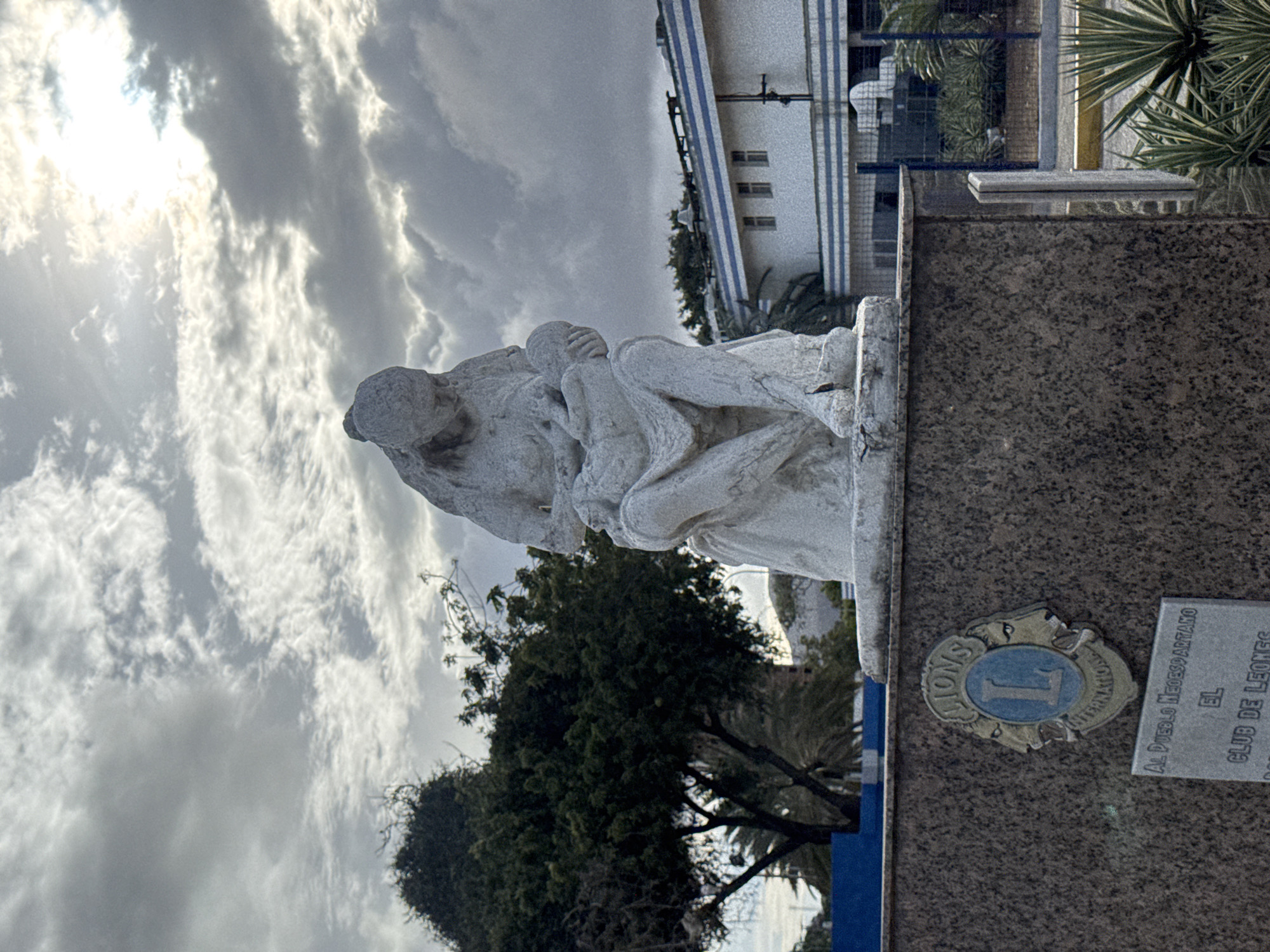 The image shows a white stone statue of a mother holding a baby, located in Porlamar, Venezuela. The statue is placed on a pedestal made of granite. A Lions Club International emblem and a plaque with Spanish text are visible on the pedestal. In the background, there is a building with blue and white stripes, vegetation, and a cloudy sky with sunlight breaking through.