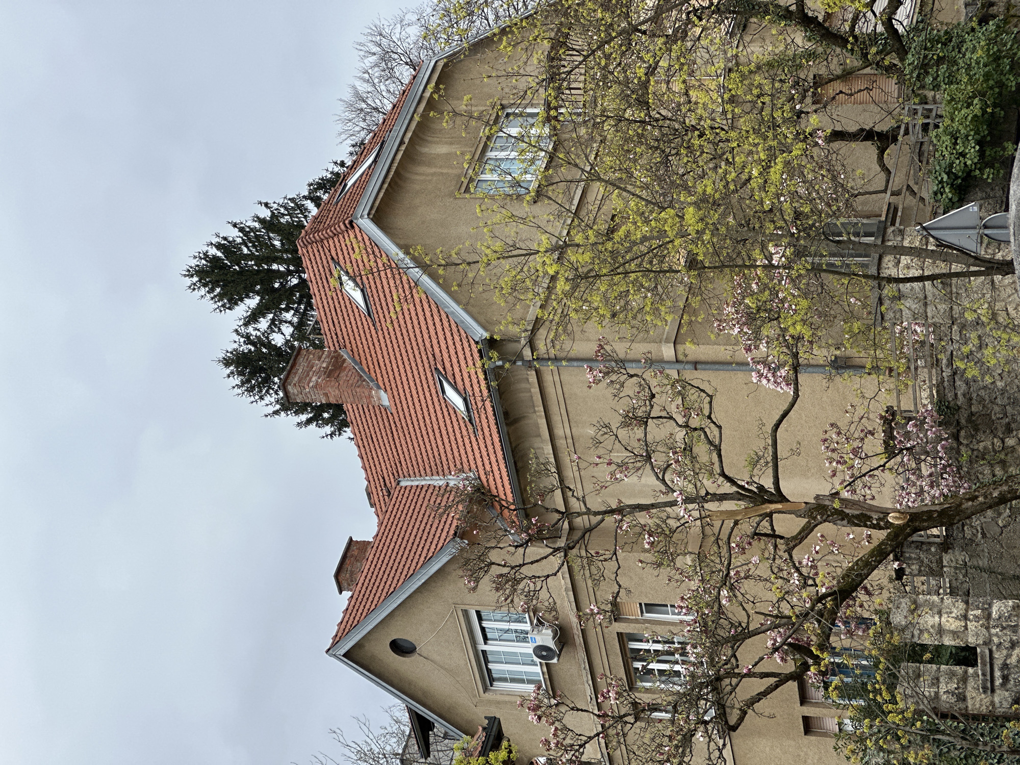 The image depicts a residential building in the City of Zagreb, Croatia, characterized by its light brown stucco exterior, red-tiled roof featuring dormer windows, and a prominent chimney. The building is partially obscured by flowering trees with pink blossoms, as well as trees with emerging yellow leaves. The structure also exhibits standard windows with white frames, an air conditioning unit installed beneath one of them, and a circular design element on its facade. The background consists of a cloudy, overcast sky. A street sign is also visible, mounted on a pole near the building.