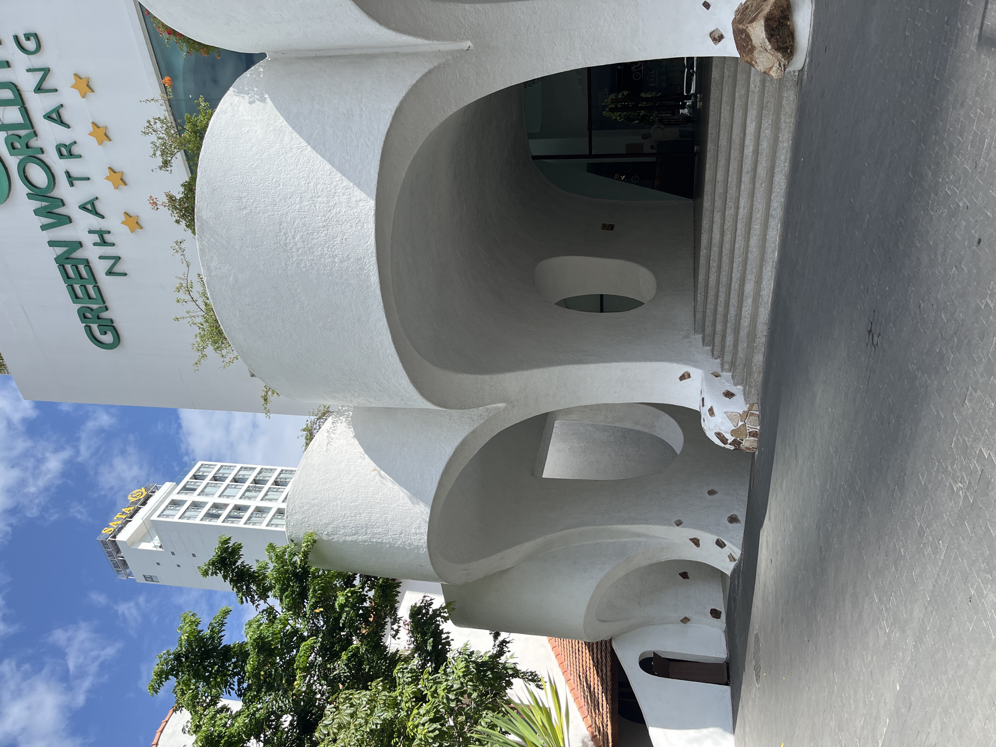 The image captures the distinctive facade of the Green World Hotel Nha Trang, Vietnam, showcasing its unique organic architectural style. The building's exterior is characterized by smooth, white, curved structures that create a series of flowing archways and recessed spaces, resembling large, smooth pebbles or abstract natural forms. The entrance is marked by a wide set of concrete stairs leading into a glass-paneled doorway.
Above the curved entryway, the hotel's name, "GREEN WORLD NHATRANG," is prominently displayed in large, green lettering against a white background. Four golden stars are arranged in a row below the text, indicating a hotel rating. To the left of the main building, a tall, white, modern hotel with the "SATA" logo visible on its rooftop is partially obscured by lush green foliage from a large tree. The foreground shows a paved driveway with a cobblestone-like pattern, leading towards the hotel. The sky above is a clear blue with scattered white clouds. The overall scene presents a striking blend of avant-garde architecture and tropical vegetation under a bright sky.