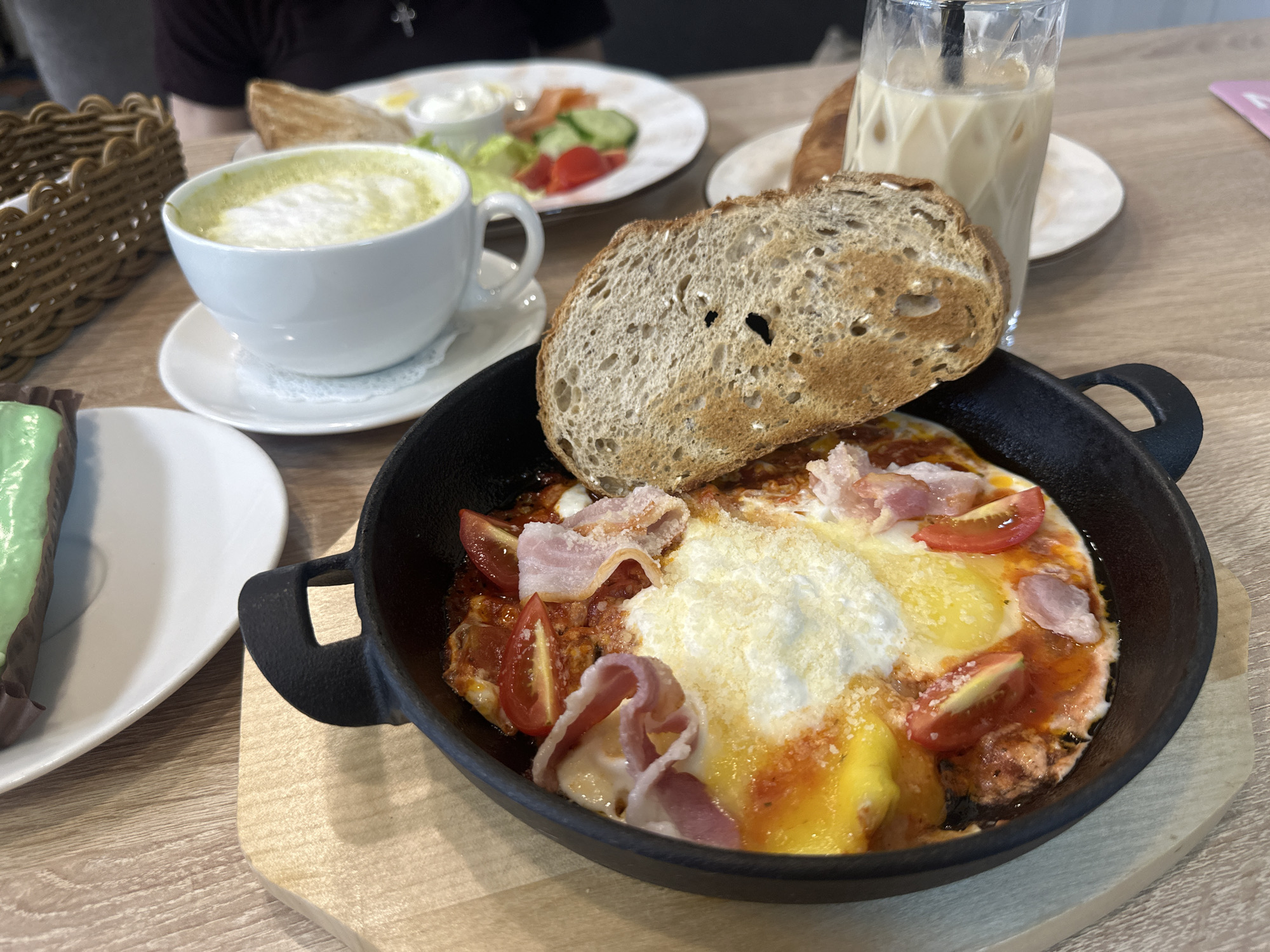 The image presents a vibrant spread of food and beverages, indicative of a breakfast or brunch setting at an indoor cafe or restaurant. The central subject is a black cast-iron skillet containing a baked egg dish. This dish features two eggs with firm whites and bright yellow yolks, nestled in a rich red tomato sauce. Halved cherry tomatoes and several strips of lightly cooked bacon are mixed into the sauce, and the entire dish is topped with a generous amount of grated white cheese. A thick slice of toasted dark bread, possibly sourdough, is leaning into the skillet. The skillet is placed on a light wooden serving board.
To the left of the skillet, a white ceramic cup holds a foamy green drink, likely a matcha latte, served on a matching saucer with a white doily. Further left, a portion of a green cake or dessert on a white plate is partially visible. In the background, a tall, clear glass filled with ice and a milky beverage, possibly an iced latte, is present. Behind this glass, part of a light-colored pastry, perhaps a croissant, can be seen. Another white plate in the background holds a small side salad with lettuce, cucumber, and tomato, alongside slices of what appears to be smoked salmon or lox, and a small white dish containing a creamy spread.
A person, wearing a dark shirt and a silver cross necklace, is partially visible in the upper left background. A brown woven basket sits on the table to the far left. The setting features a light wood-grained tabletop, suggesting a bright and casual atmosphere suitable for a daytime meal. There is no visible text in the image.