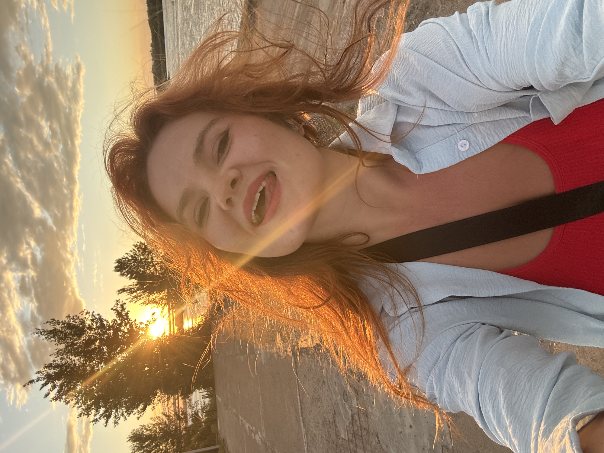 A young woman with long, red hair is taking a selfie outdoors during sunset. She is wearing a light blue button-up shirt over a red top. Her mouth is open as if she is speaking or laughing, and she has a bright smile. The setting appears to be a concrete embankment next to a body of water, likely a river or lake. Trees are visible in the background, silhouetted against the warm glow of the setting sun. The sky is filled with dramatic clouds, illuminated by the golden hour light, which also casts long shadows and highlights the texture of the concrete. The overall mood is joyful and vibrant, capturing a moment of happiness during a beautiful sunset in Nizhny Novgorod, Russia.