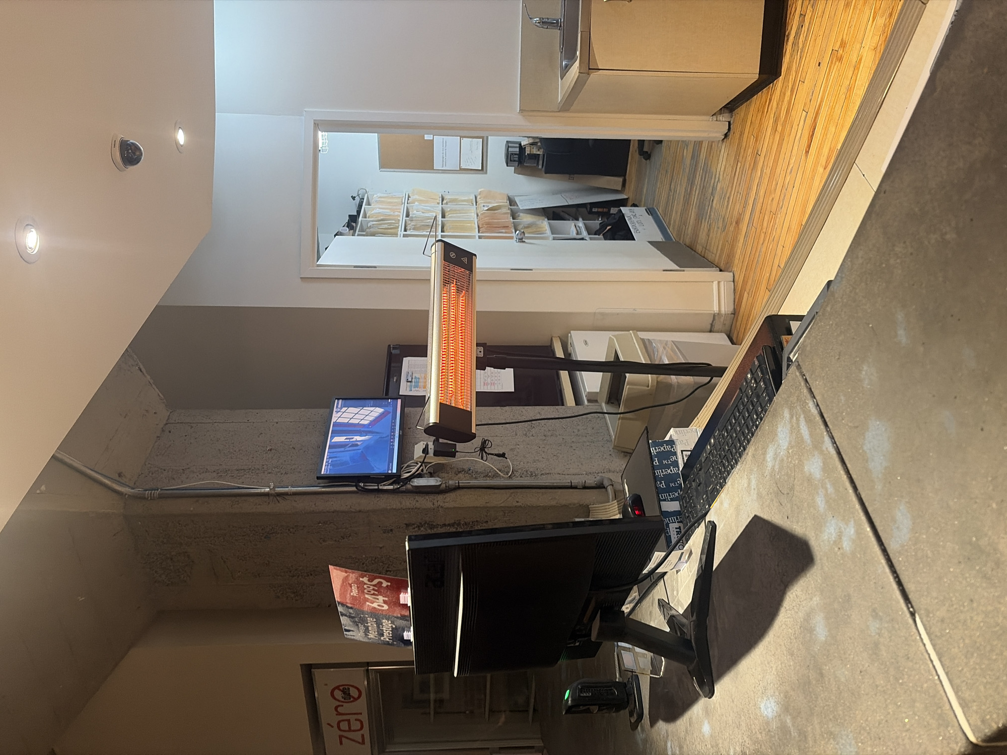 This media file captures an indoor commercial or office environment, specifically a reception or workstation area with a view into an adjacent office space. The scene is illuminated by artificial lights, including recessed ceiling lights and the glowing element of an electric heater, suggesting it is either indoors during the day or evening.
In the foreground, a large, dark-colored monitor sits on a concrete-textured countertop, accompanied by a keyboard and mouse. A bright orange electric infrared heater is prominently displayed above this desk area, mounted on a pole or stand. To the left of the heater, a smaller monitor displays what appears to be a 3D rendering or architectural design. Cables and metallic conduits run along the textured concrete wall behind these monitors. A white wall to the left features two recessed lights and a dark-domed security camera.
Through an open doorway or window in the background, another room is visible. This room features light-colored wooden flooring and shelves filled with numerous organized folders or files, suggesting a back-office or record-keeping area. A small sink and countertop are also visible in this adjacent space, along with what appears to be a corkboard holding papers. Office equipment, possibly a printer or scanner, can be seen near the shelves.
No people are present, and no active event is taking place, giving the scene a quiet, unoccupied atmosphere.
Visible text includes:
* On a red sign or poster near the foreground monitor: "Promo", "Membres", "649$", "Prestige", and "Destine".
* On the base of one of the monitors: "zéro".
* Partially visible on a box or sign near the keyboard: "Paperin".
* On the heater itself, some symbols (possibly warning or operational icons).