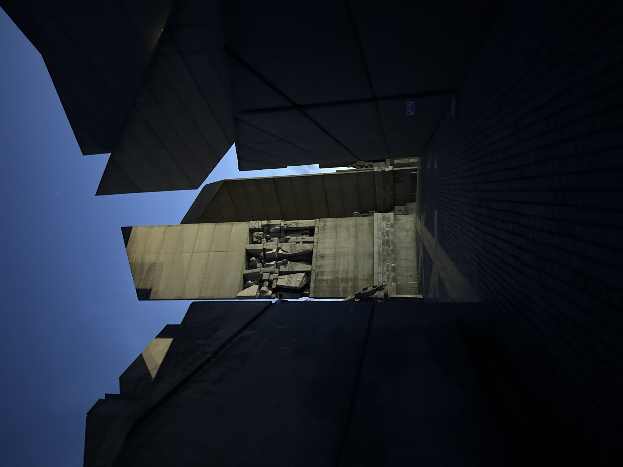 The image shows a close-up, low-angle shot of the Founders of the Bulgarian State Monument in Shumen, Bulgaria. The monument features large, angular concrete blocks that create a dramatic and imposing structure. A blue sky is visible in the spaces between the blocks. The monument's relief, with carved figures, can be seen on a vertical block. The angle of the shot emphasizes the monument's height and scale. The lower portion of the image is mostly dark, contrasting with the lighter, sky-exposed elements.