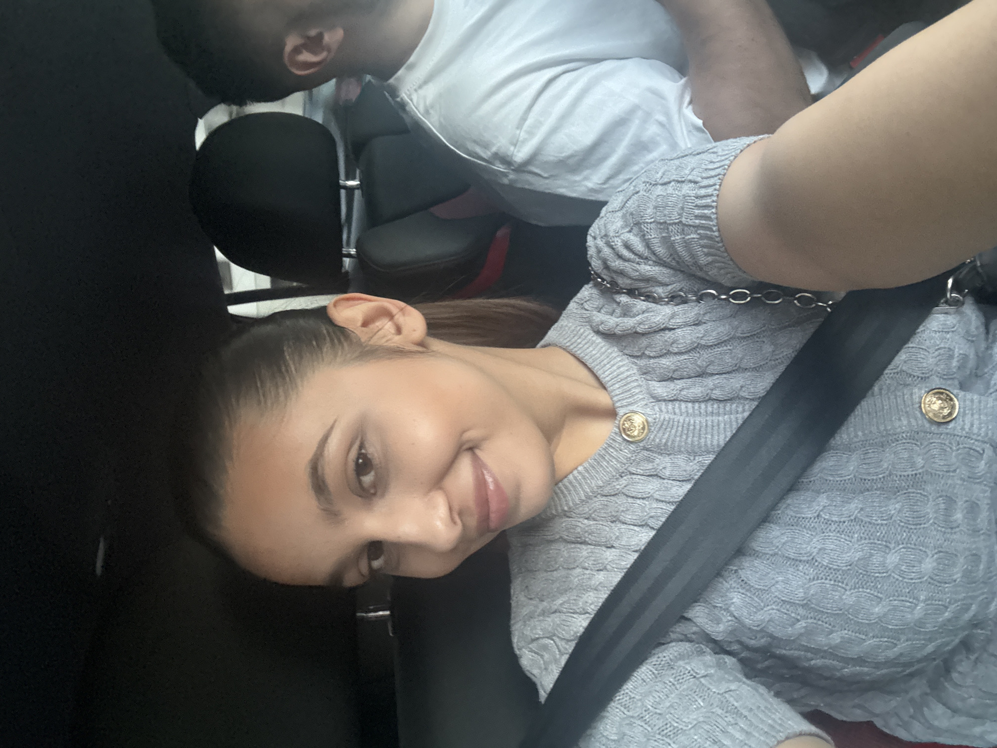 A young woman wearing a gray sweater with gold buttons and a seatbelt across her chest is in the foreground. A man in a white shirt is visible in the background, seated in a car. The car's interior is visible, including black seats. The specific location in Mönchengladbach, Germany cannot be determined from the visual information provided.