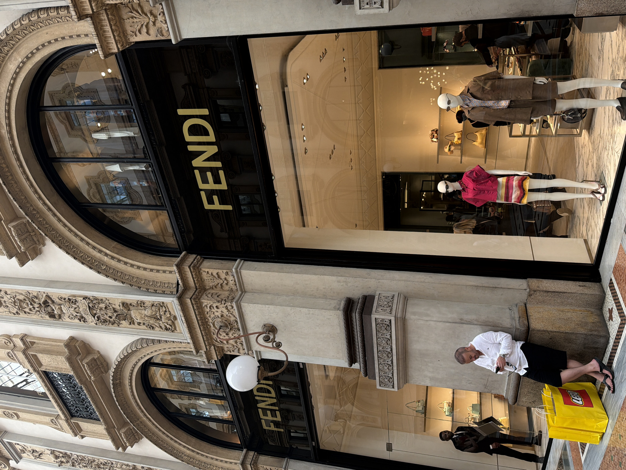 This image shows the exterior of a Fendi store in Milan, Italy. The building has ornate architectural details, including arched windows and carved stonework. The Fendi sign is prominently displayed above the storefront. Inside the store's windows, there are mannequins wearing designer clothing. In the foreground, a man is sitting next to a bright yellow LEGO bag. Another man in a suit is reflected in the storefront glass. The scene captures a blend of high fashion and street-level activity in a historic urban setting.