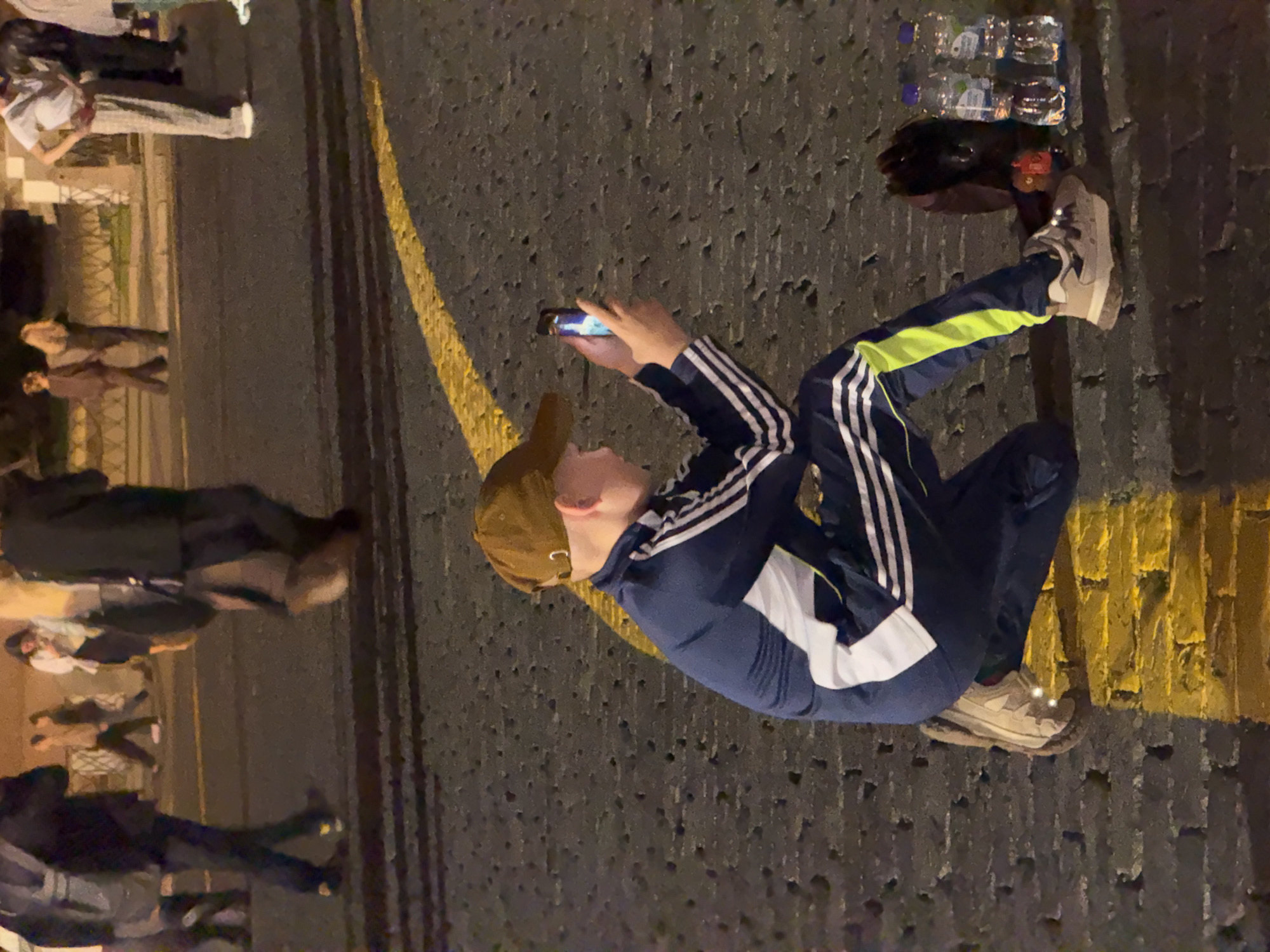 The image shows a young person with a brown baseball cap, wearing a navy blue tracksuit with white and yellow stripes, kneeling on a cobblestone street with a yellow line painted on it. The person is holding a smartphone and appears to be taking a photo or video. In the background, several other people are walking or standing. The scene is illuminated by artificial light, suggesting it is nighttime or dusk. The environment appears to be a public area, possibly a street or square. The style of the tracksuit and the presence of multiple water bottles near the kneeling person suggest a casual setting. The presence of the yellow line on the cobblestones could indicate a pedestrian crossing or a designated path. Without further context or identifying features, it is impossible to determine the specific location in Moscow.