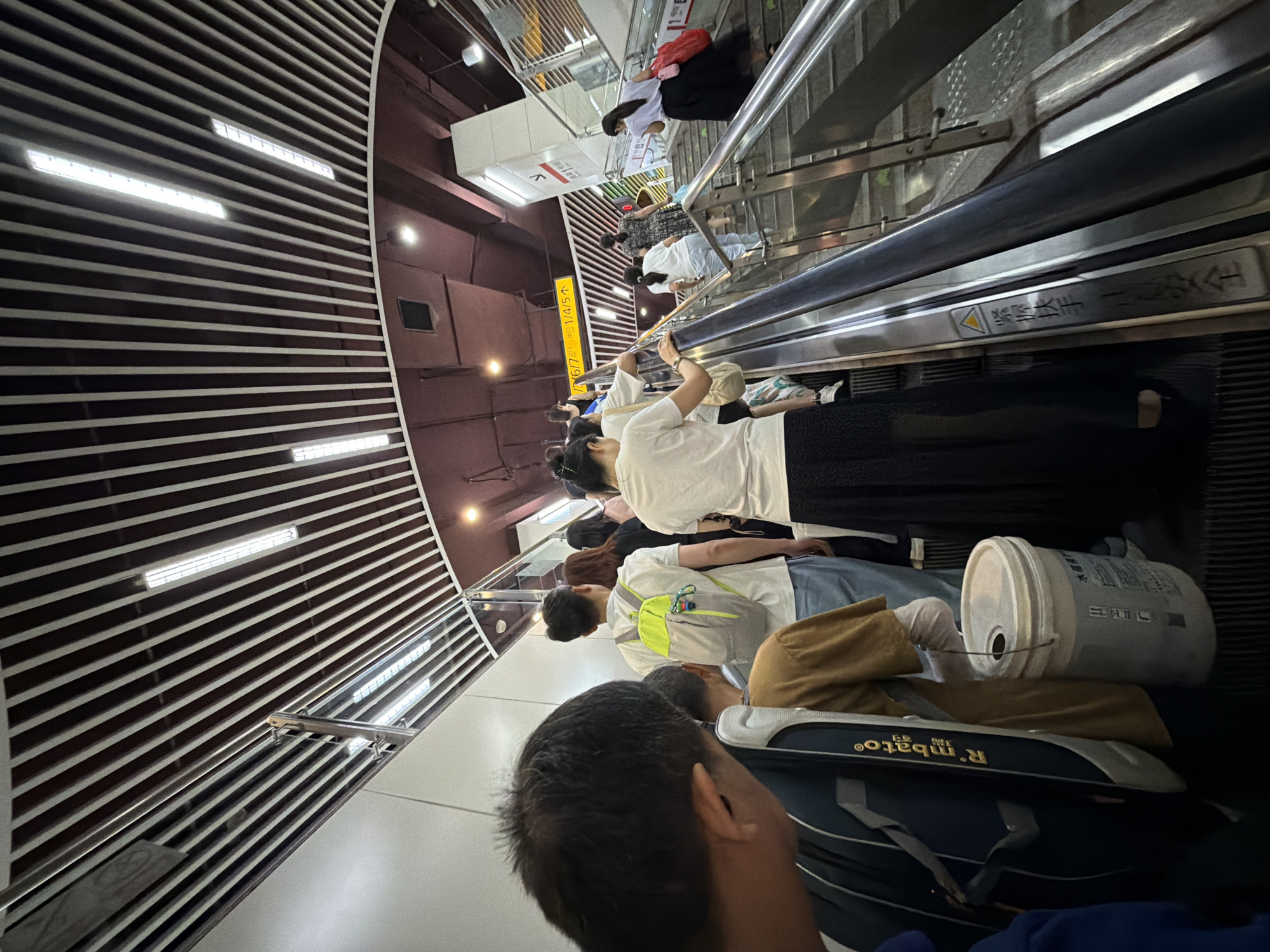 In the Jiulongpo District, China, a group of people are seen ascending an escalator. The scene appears to be within a public transport hub or a similar large building, characterized by a modern, slatted ceiling with integrated lighting. On the escalator, individuals are dressed in casual attire. One person wears a white t-shirt and a long black skirt, holding a red bag. Another person has a light-colored backpack with a bright green accent. A man in the foreground is partially visible, with the back of his head facing the camera and carrying a dark backpack. Further up the escalator, more individuals are ascending, including a person in a patterned dress. The escalator itself is metallic with safety railings and cautionary signage.
