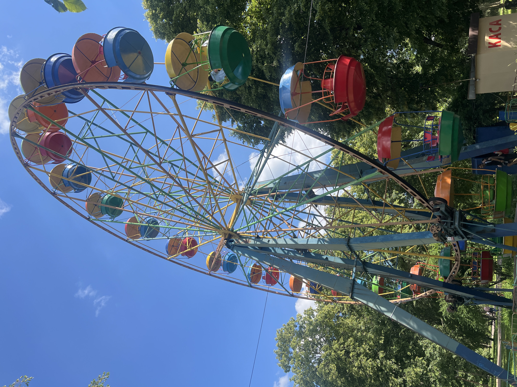 A brightly colored Ferris wheel stands in a park on a clear day. The Ferris wheel has many gondolas of various colors, including red, blue, green, and yellow. The wheel is supported by a large, multi-colored metal structure. In the background, lush green trees form a dense canopy, suggesting a warm, sunny environment. Sunlight filters through the leaves, casting shadows on the ground. A small building with the word "KACA" in large red letters is visible near the base of the Ferris wheel. A few people can be seen near the base of the Ferris wheel, appearing to be interacting with the attraction or its surroundings. The overall scene depicts a lively and recreational atmosphere within a park setting.