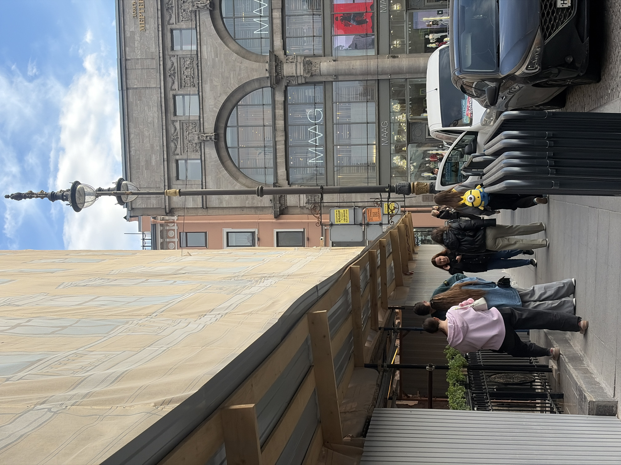 The image captures a street scene in Saint Petersburg, Russia, featuring pedestrians walking along a sidewalk next to a building under renovation, as scaffolding covered with protective material obscures part of the view. A streetlamp stands prominently on the left.
On the right, pedestrians are visible, including an individual wearing a pink sweatshirt and another carrying a yellow Minion plush toy. Adjacent to the pedestrians are parked cars.
Across the street is a large building with the name "MAAG" displayed prominently in the windows. The building is of classical architectural style with large arched windows. The sky is partially cloudy.