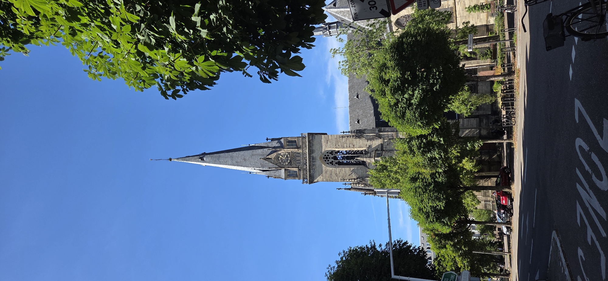 The image depicts a view of the exterior of the Cathédrale Notre-Dame de Strasbourg, France, on a clear, sunny day. The prominent feature is the cathedral's spire, a tall, pointed structure made of grey stone, reaching towards a bright blue sky. The spire is adorned with Gothic architectural details, including smaller turrets and decorative elements. A clock is visible on the tower section of the spire, showing the time.
In the foreground, a street with road markings is visible, including white lines and the word "ZONE" painted on the asphalt. Several cars are parked along the side of the street, and a bicycle rack with parked bicycles is also present. Lush green trees with dense foliage are scattered throughout the scene, partially obscuring the lower parts of the cathedral and providing shade.
Further to the right, a street sign with French text and a speed limit of "30" is visible, indicating a traffic regulation zone. The overall environment suggests an urban setting, likely a public space near the cathedral. No people are clearly identifiable in the image, though their presence is implied by the parked vehicles and bicycles. The lighting suggests it is daytime, with sunlight casting shadows.