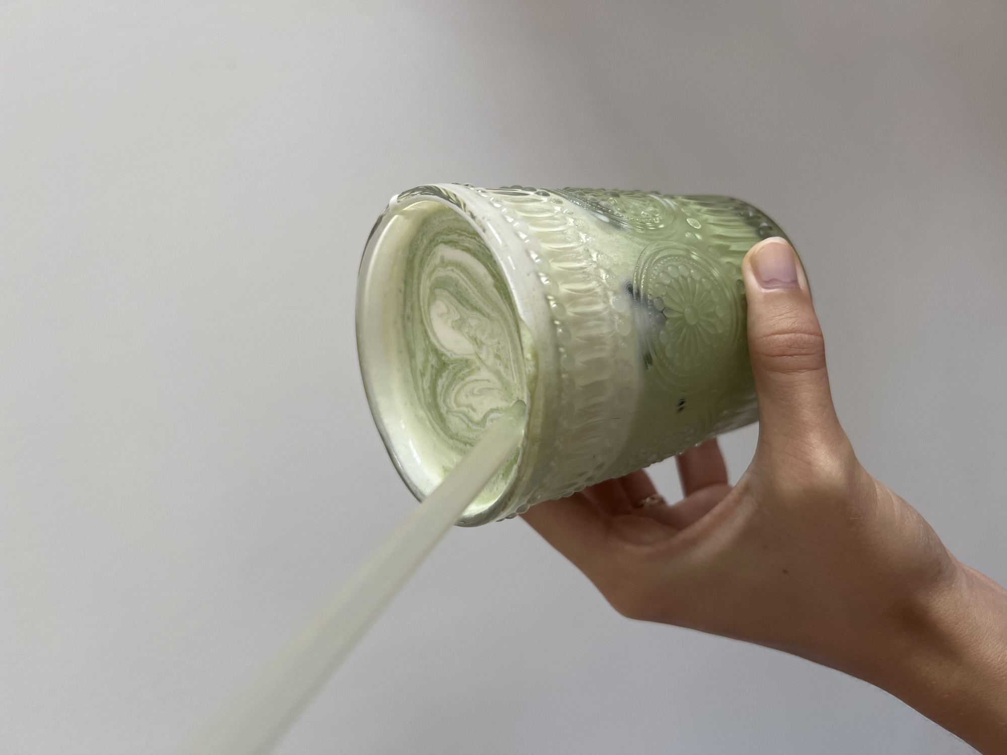 A hand is holding a glass with a green and white layered beverage, likely matcha latte. The glass is tilted, and a liquid is being poured from it. The background is a plain grey wall. The location cannot be determined based on the image.