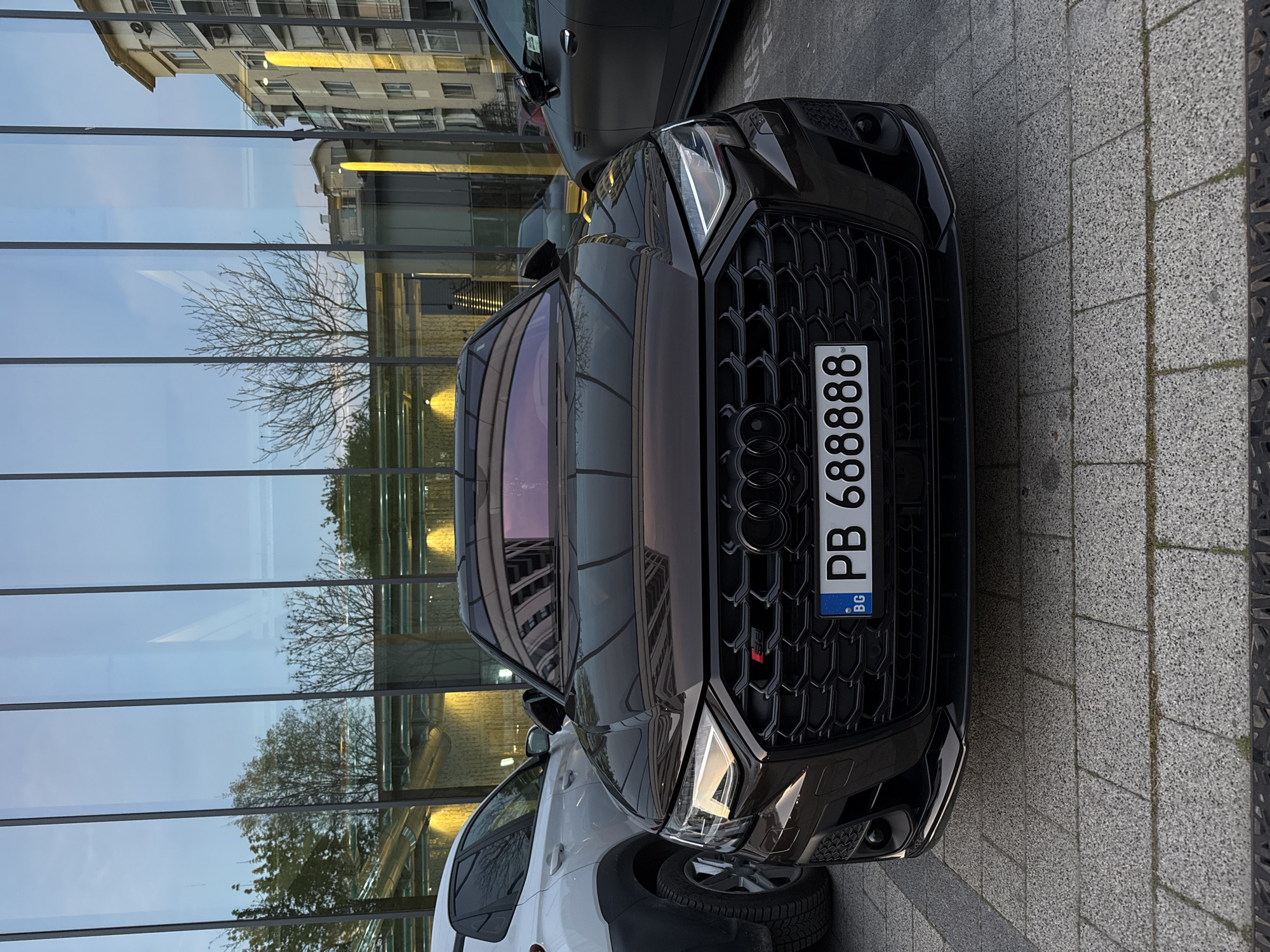 The image shows the front of a black Audi RS5 parked on a paved area. The car has a Bulgarian license plate that reads "PB 6888BB". The background features a modern building with large glass windows that reflect the surrounding cityscape. The building's design suggests an urban environment. The location is consistent with the description of Plovdiv, Bulgaria, given the license plate and architectural style.