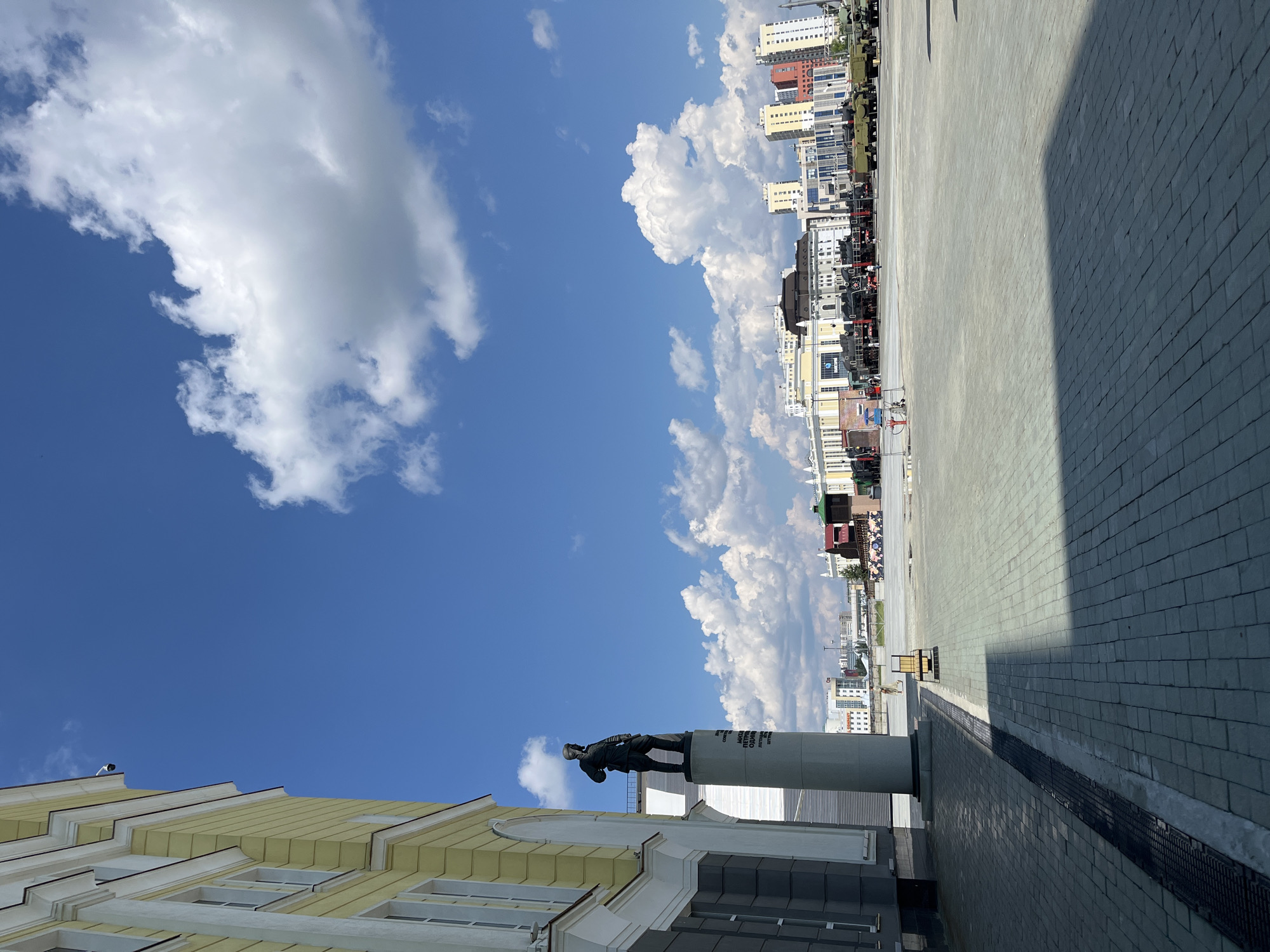 The image depicts a wide, paved plaza in front of a yellow and grey building, likely a museum or historical site, in Verkhnyaya Pyshma, Russia. A prominent statue of a man in military attire stands on a tall cylindrical pedestal to the left of the frame. In the background, a row of buildings, including multi-story structures and what appears to be a museum exhibiting trains and military vehicles, stretches across the horizon. The sky is a vibrant blue with scattered white cumulus clouds. A few individuals are visible in the distance, walking across the plaza. The scene is bathed in bright sunlight, casting shadows across the paved ground.