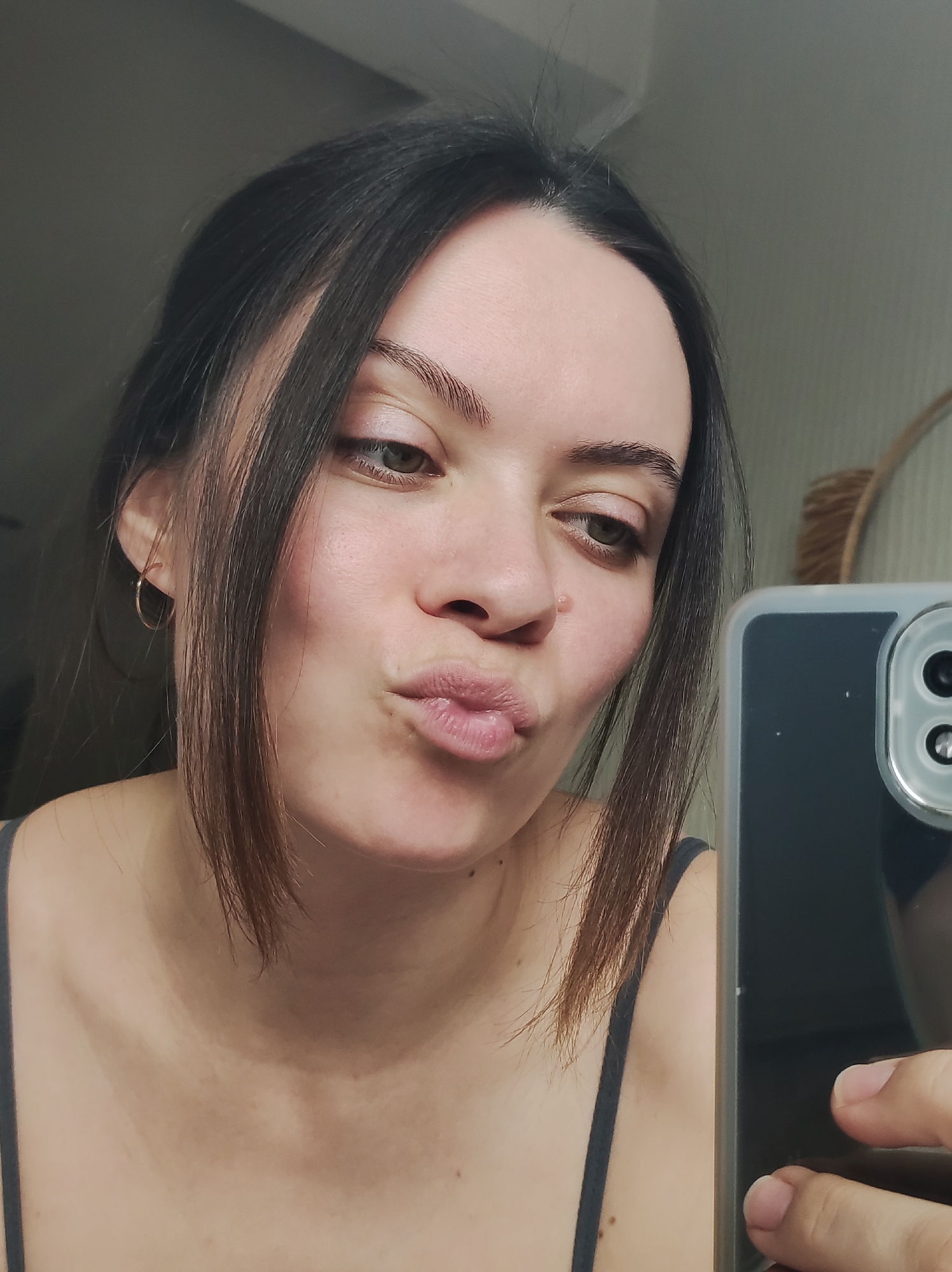A young woman with dark hair partially pulled back is taking a selfie. She has fair skin, dark eyebrows, and green eyes. Her lips are puckered in a kiss. She is wearing a gray tank top and small hoop earrings. A phone is visible in the lower right corner of the frame, held in her right hand. The background is blurred, but a light-colored wall and a partially visible round object are discernible. The location is indeterminate.